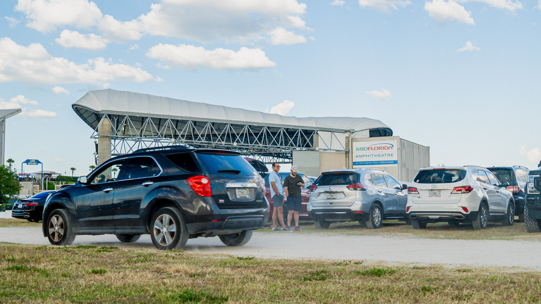 A car drives past a row of parked cars, with two fans standing nearby. MidFlorida venue visible in the background of the parked cars. 