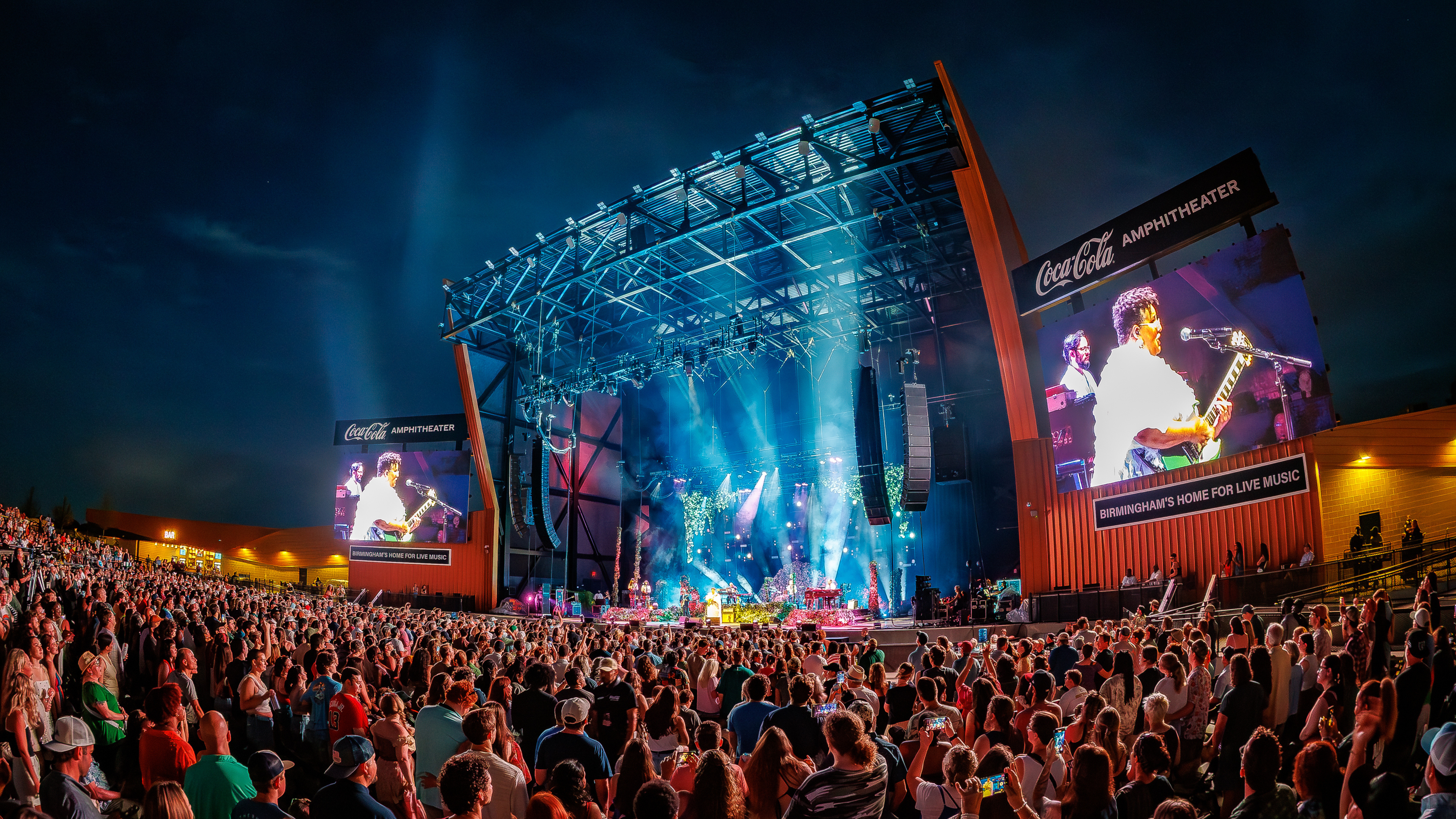 A nighttime concert at a packed outdoor amphitheater with colorful stage lights, large video screens, and a crowd facing the stage.
