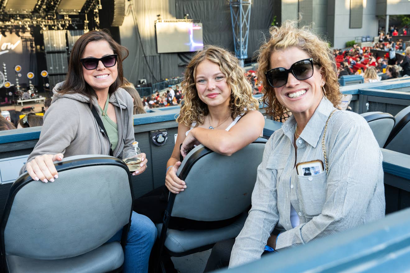 3 women sitting in premium box seating inside the venue, one with a glass of white wine, while they wait for the concert to start.