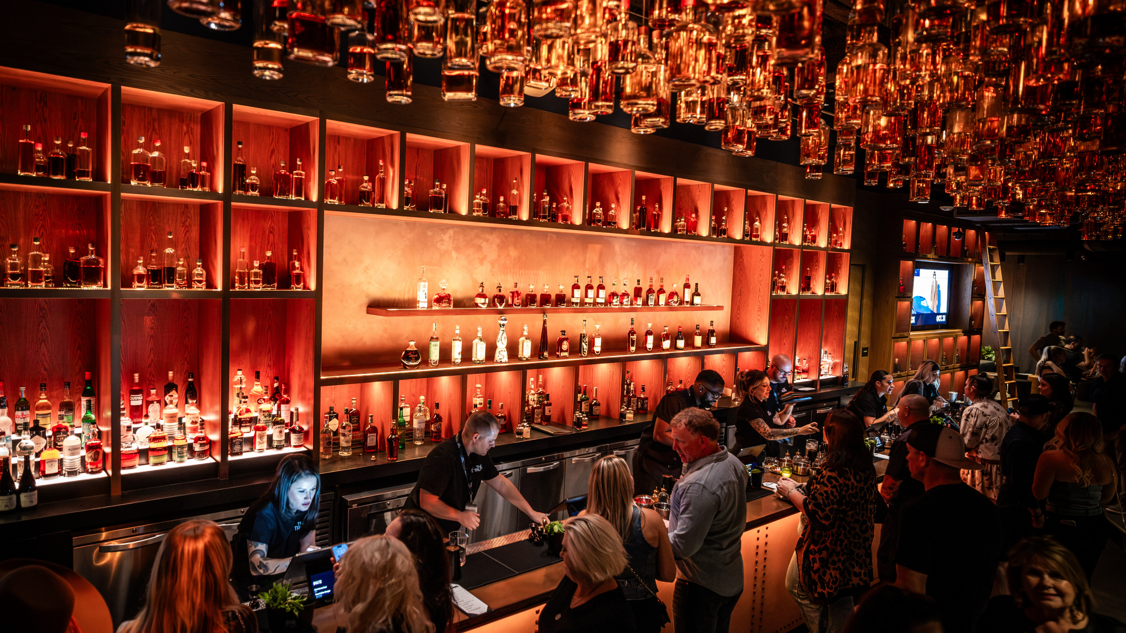 Guests gather at a lively bar beneath an installation of suspended bottles, with illuminated shelves of spirits behind bartenders. Photo credit: Alive Coverage