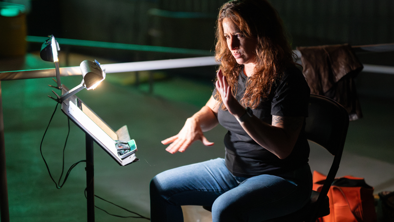 A sign language interpreter using signed language while sitting at table with a book and a table lamp in a dimly lit setting.