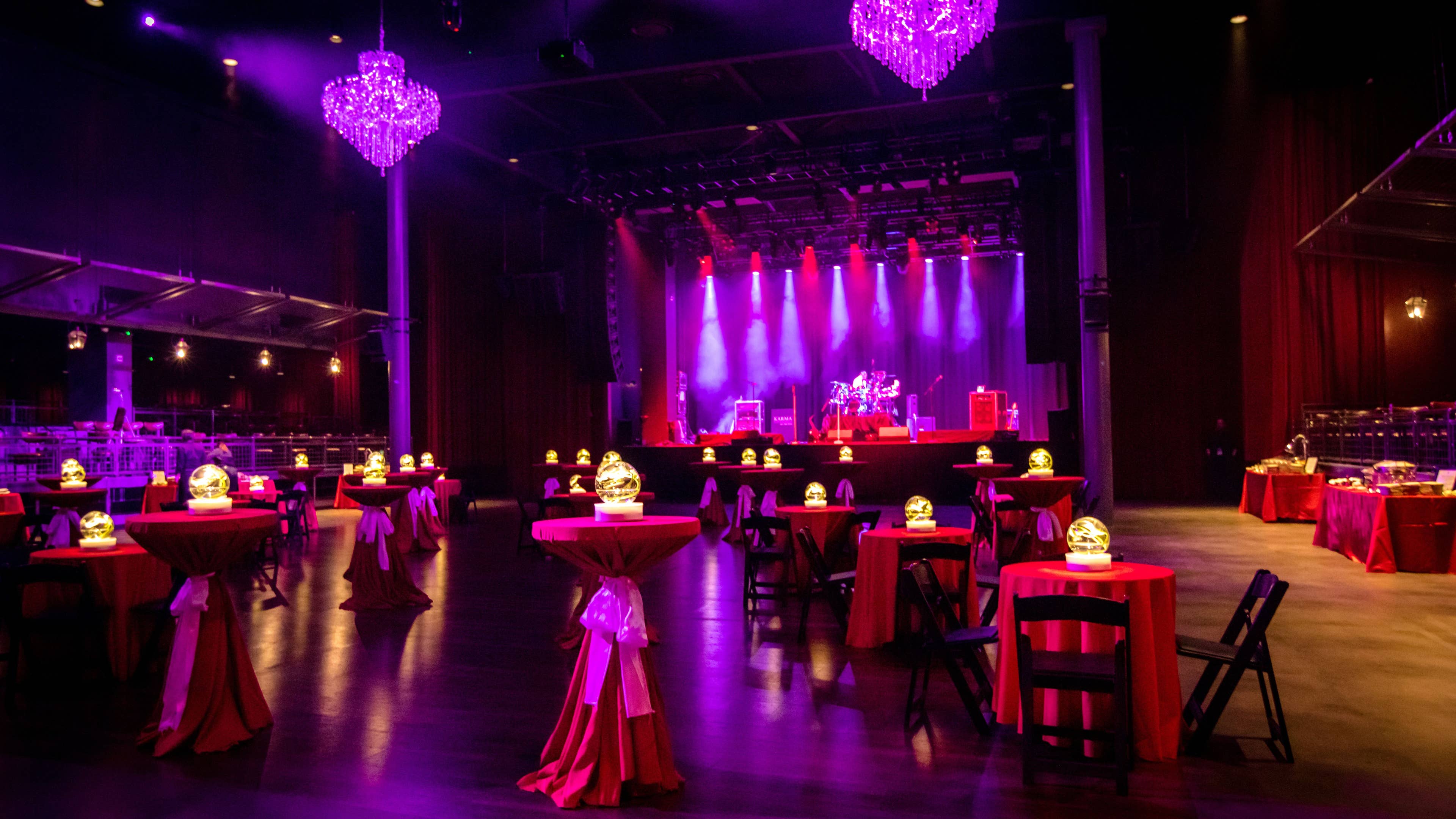The main floor of The Fillmore New Orleans decorated with chandeliers, ambient lighting, themed decor, dining tables with seating, a food station, and a band on stage