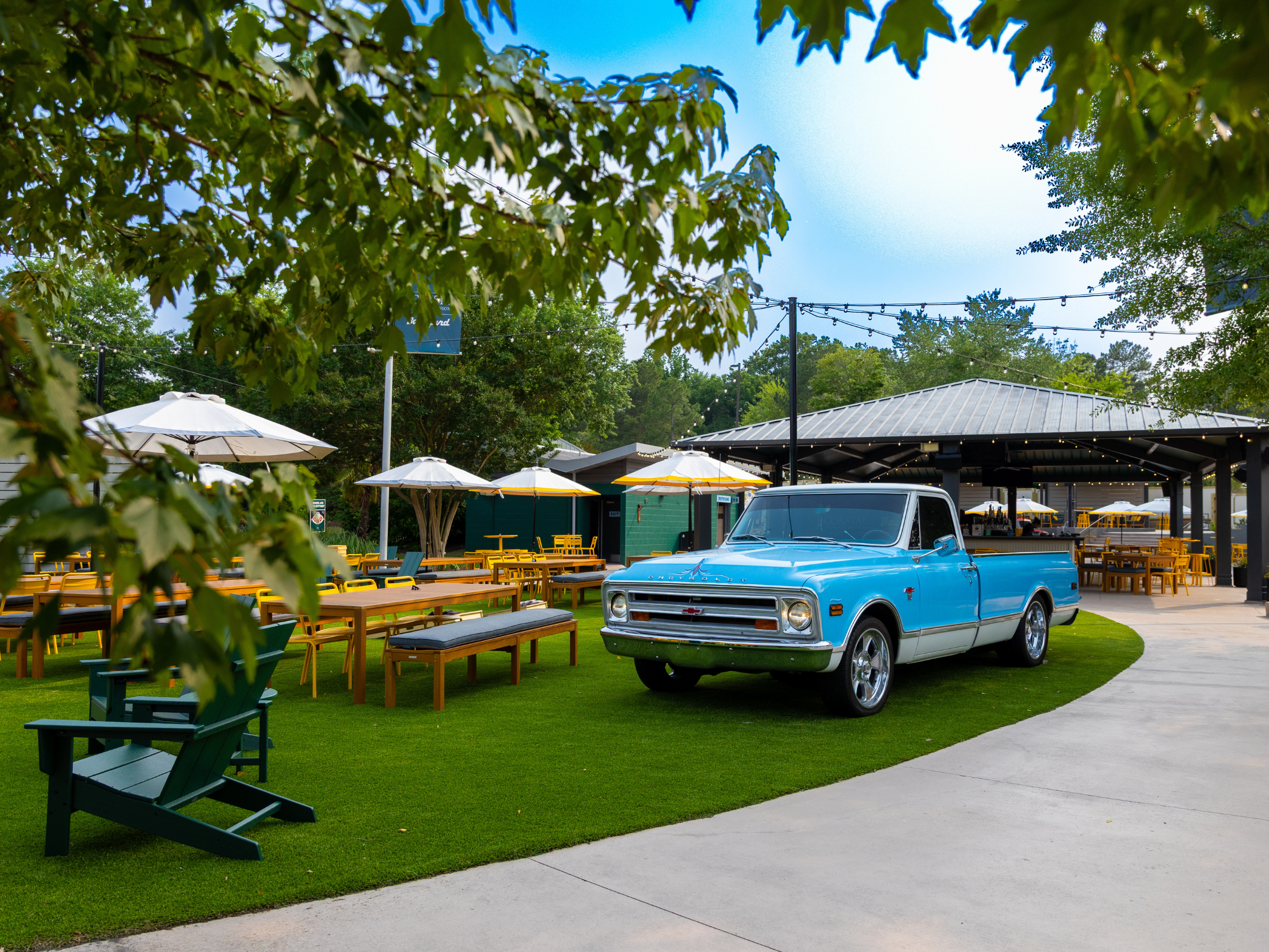 An outdoor space with tables, benches, umbrellas with a parked truck on turf. 