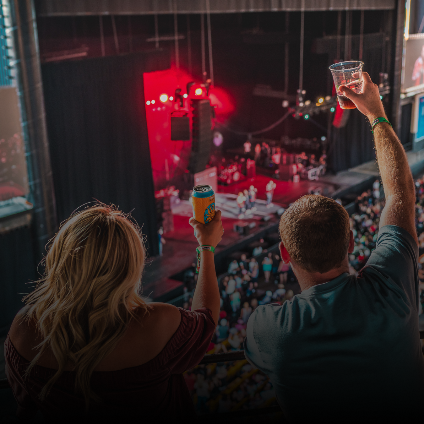 A man and a woman saluting "Cheers" with their drinks in the air while watching an artist on stage. 