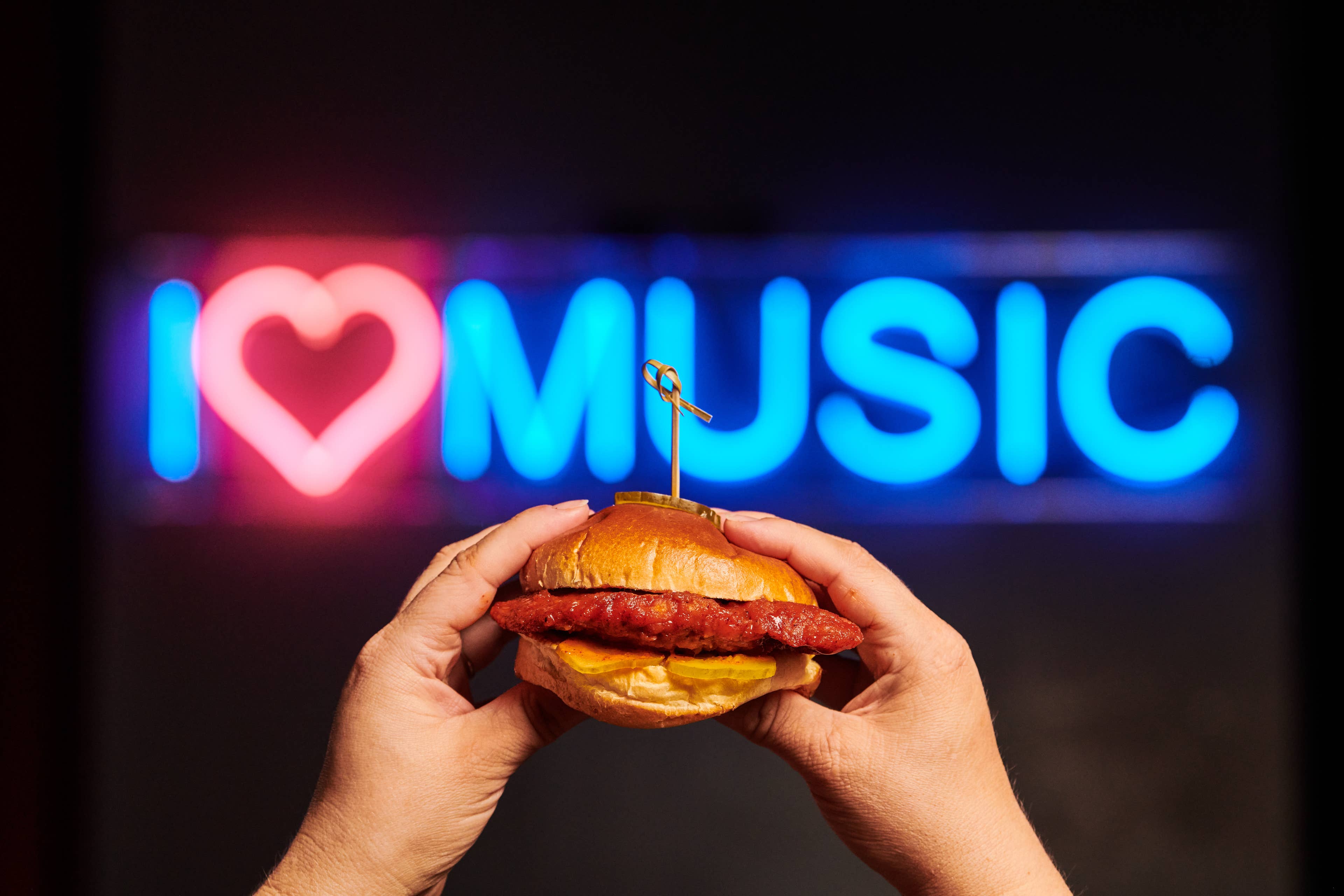A person holding a chicken sandwich in front of a sign that says "I love music".