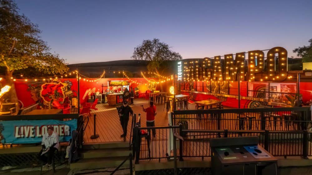 A far away shot of Mi Campo Deck, showing the outdoor patio in the evening, lit up by string lights. Tables and chairs and neon lights and art are on the walls, as well as a bar with marquee letters that spell out MI CAMPO.