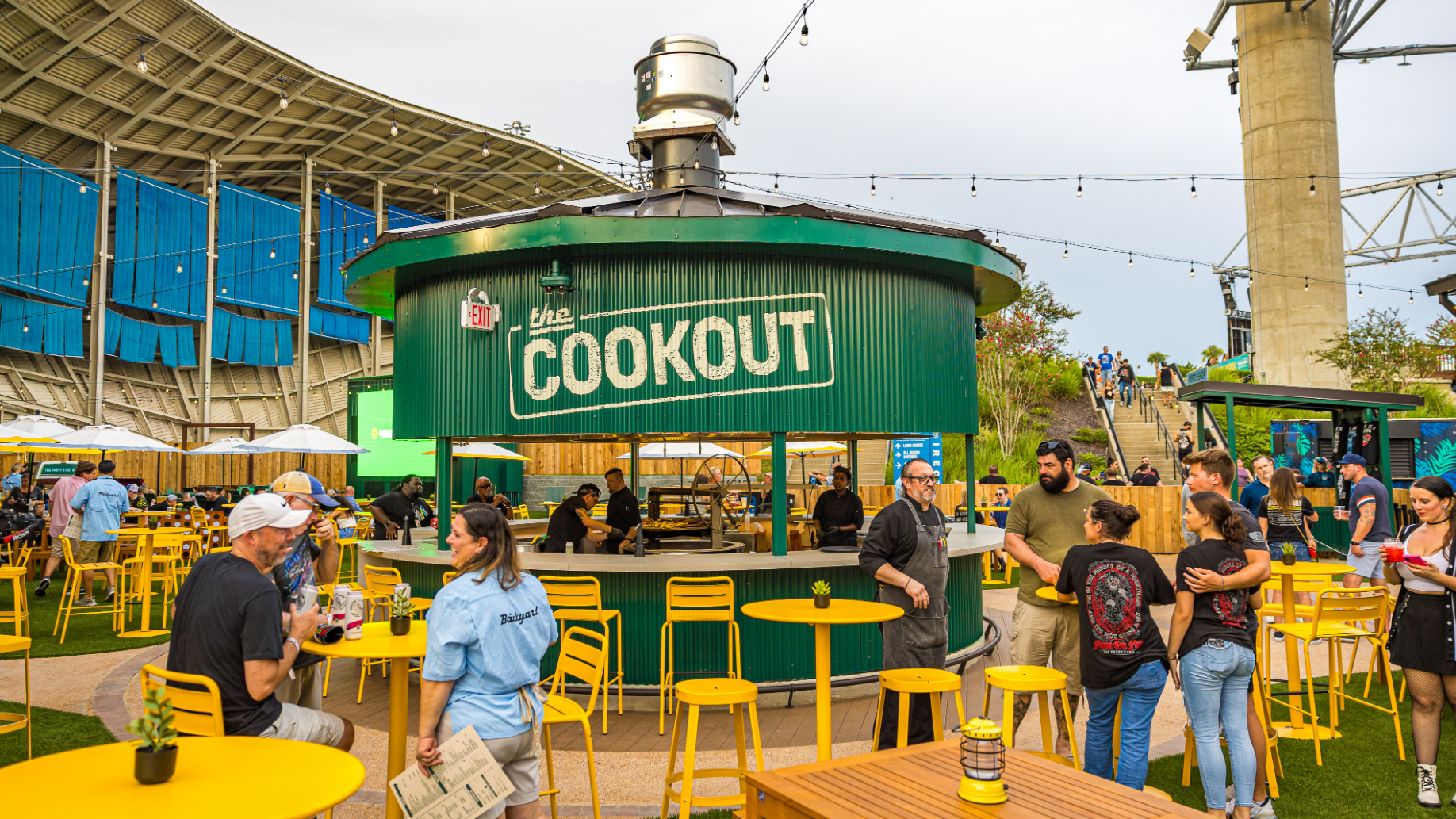 Fans sitting and standing around yellow high-top tables and chairs placed in grass. Large green cookout in the center with employees grilling. Dangling lights strung across the entire space.