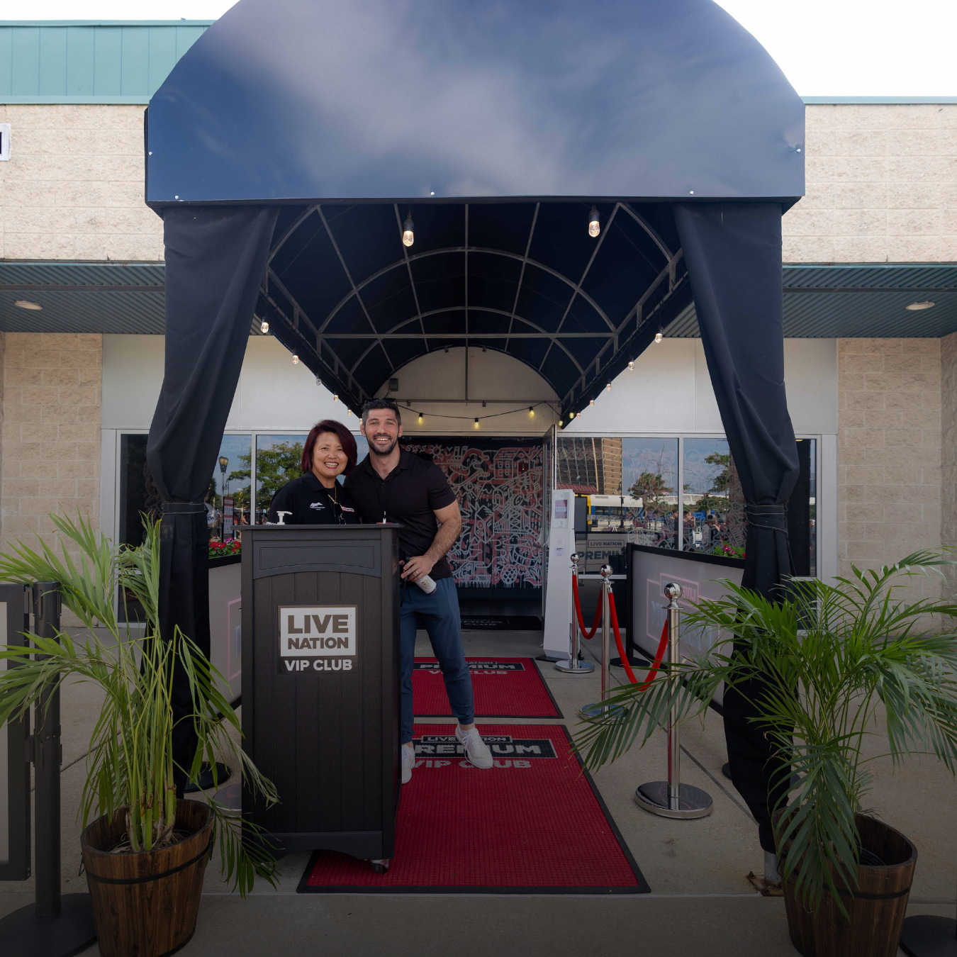 Two VIP employees smiling for the camera at the entrance of the VIP club (a black awning, black podium, and red carpet at the entrance)