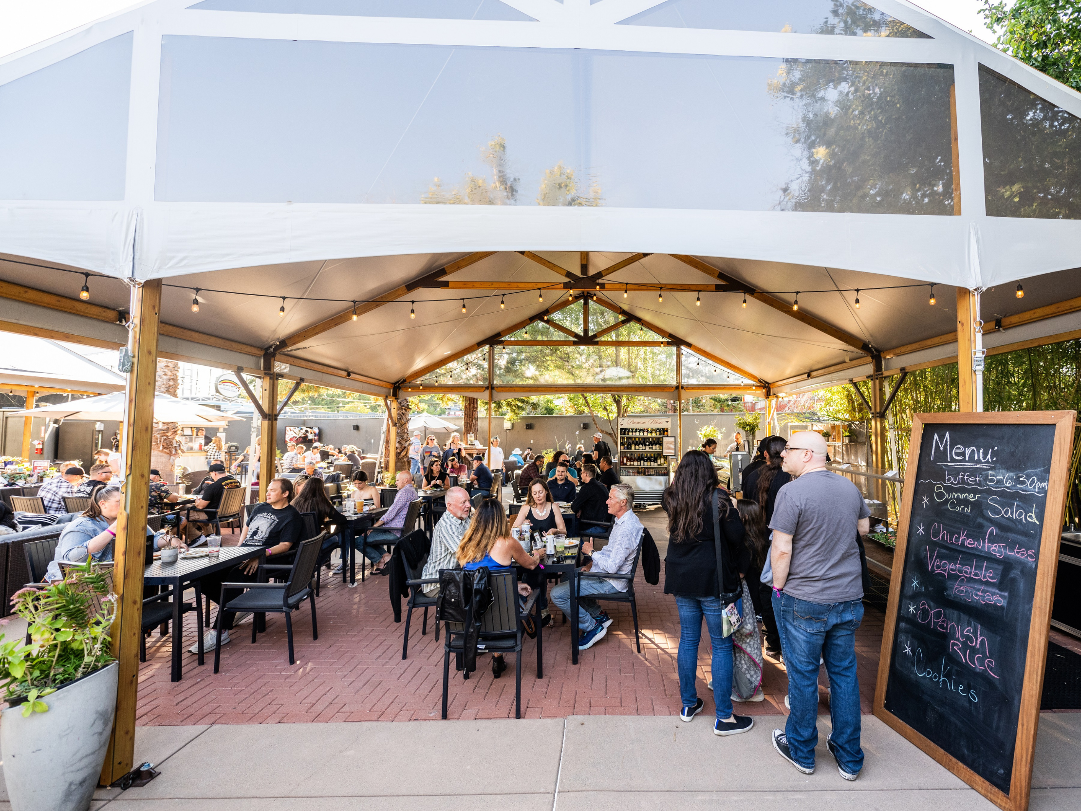 VIP Club at Shoreline Amphitheatre with people seated at tables eating and drinking under a tent with string lights, and standing in line at a buffet