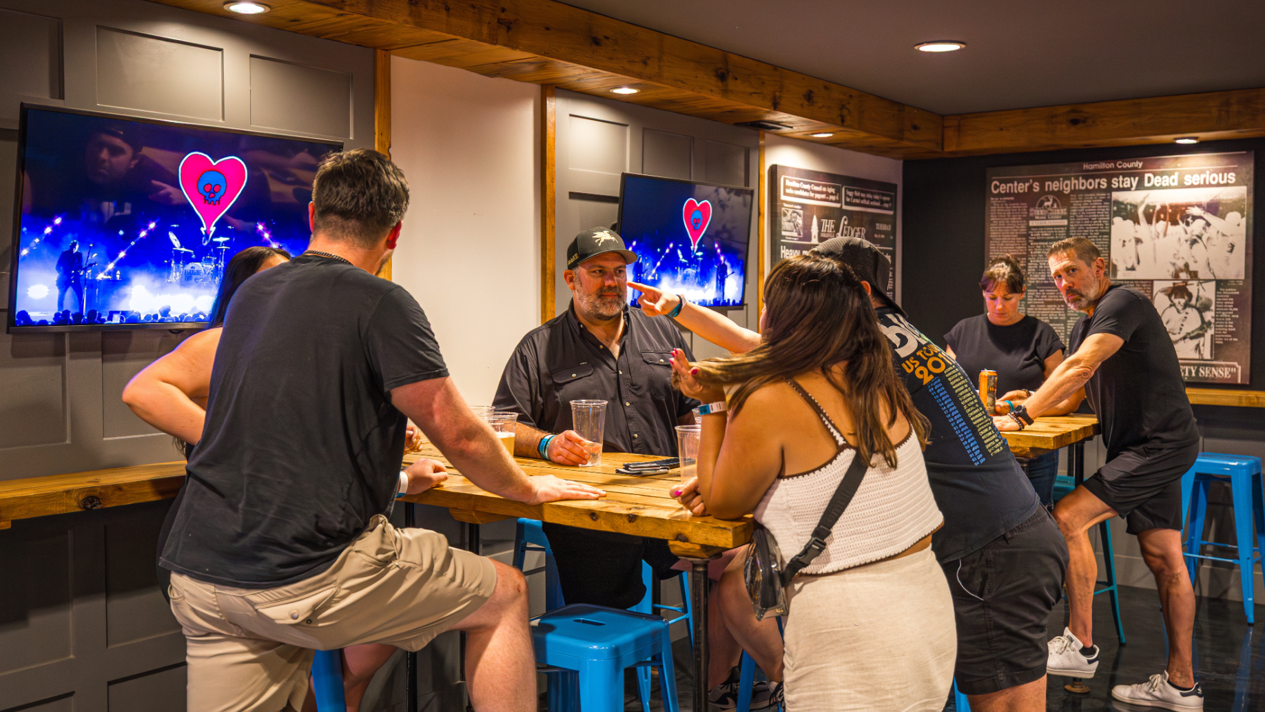 Fans sit around a high-top table chatting and having drinks while watching a live show feed on various television screens. 