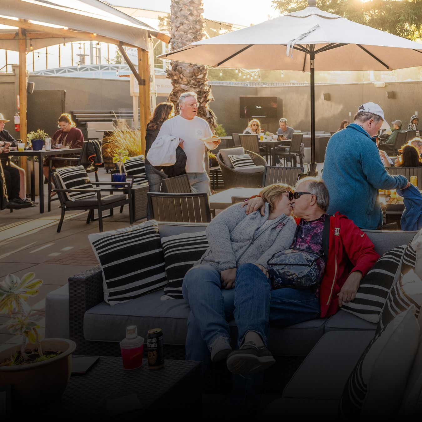 A couple embracing each other on a couch seated in the VIP area surrounded by tables, umbrellas and other fans