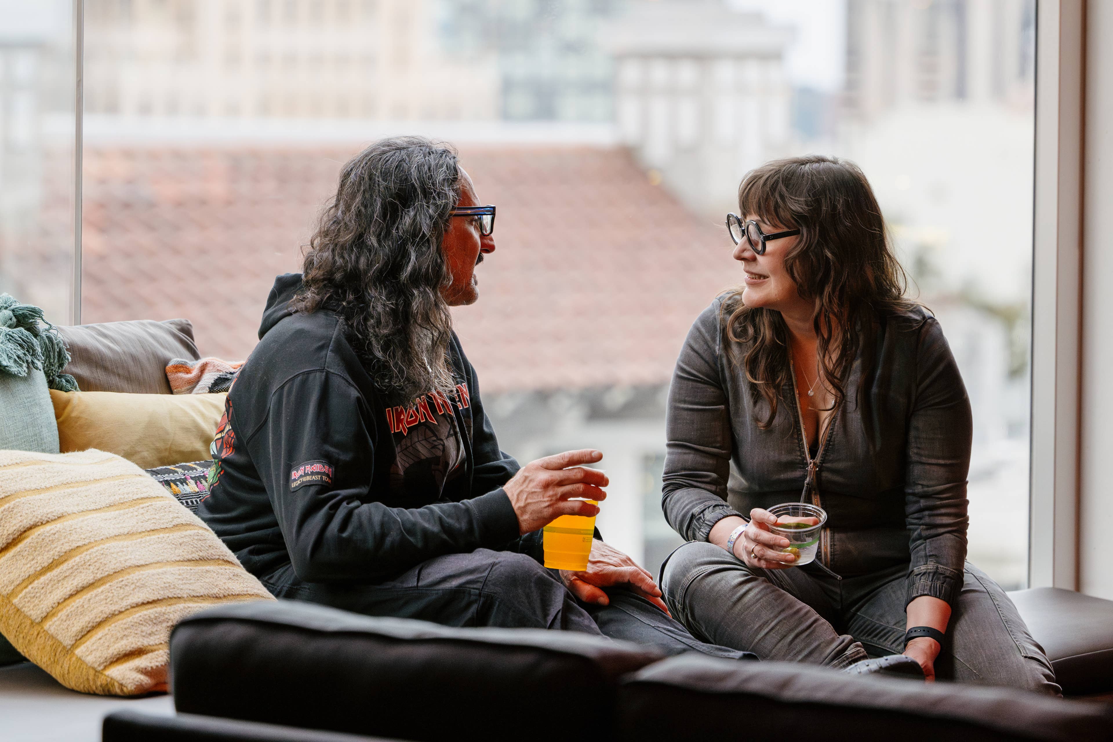 Two women sit on a cushion in front of a large window talking to each other and holding drinks.