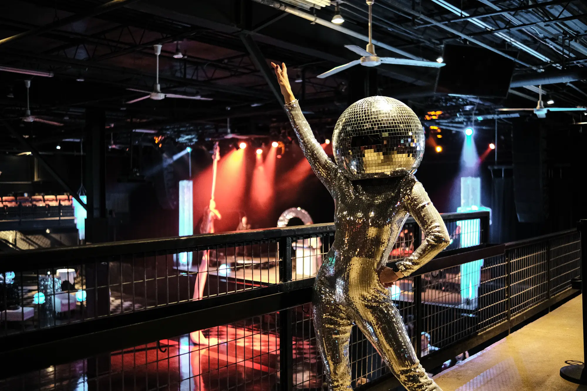 A woman in a silver sequin dress and disco ball head stands on a balcony at a concert venue