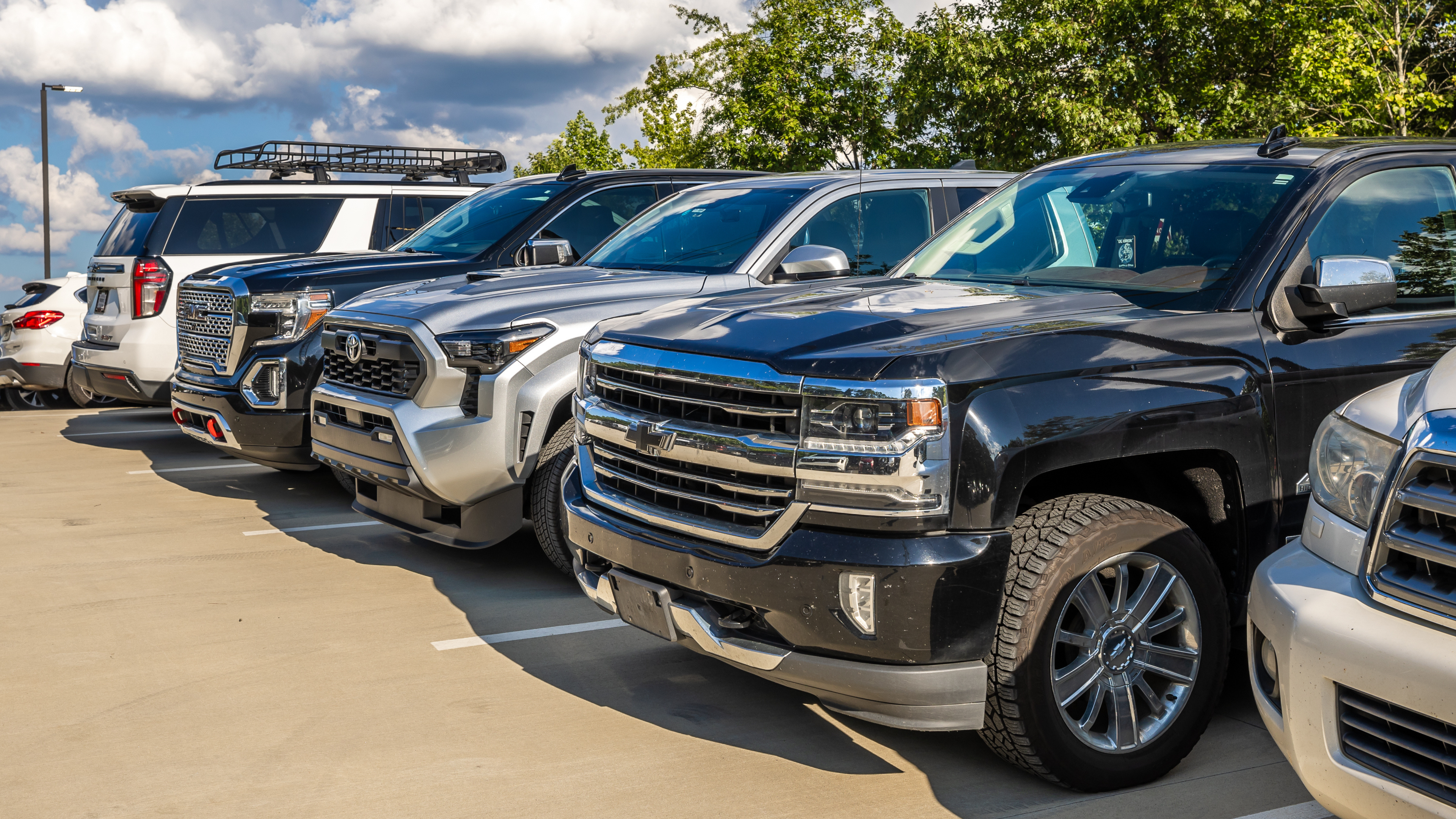 Row of parked SUVs and trucks in a parking lot across from a venue entry gate on a sunny day, lined up in marked spaces with trees in the background.