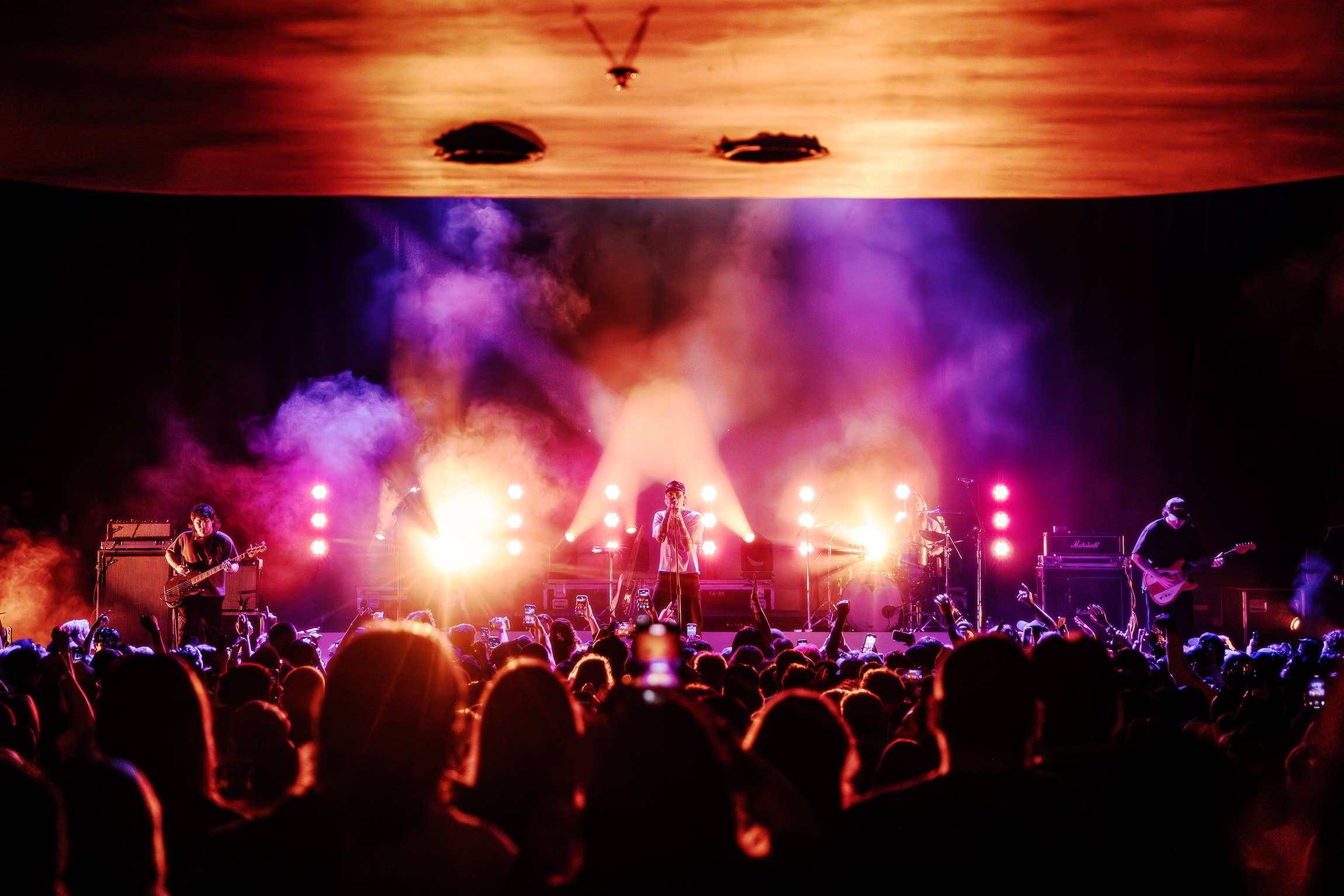 Fans in the foreground watching an artist perform onstage surrounded by purple, pink and orange lights and smoke.