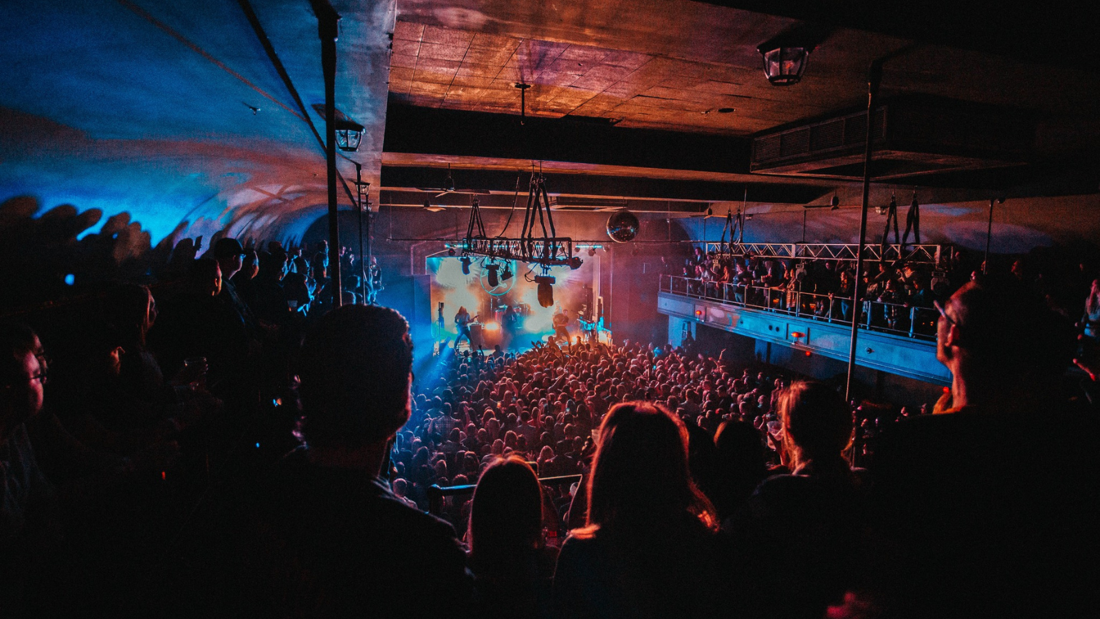 A view from the back of the venue looking towards the stage with a large crowd and show lighting. 