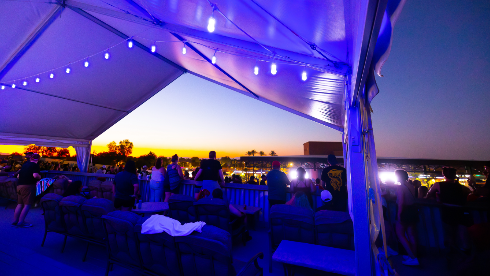 The Oasis Lounge at Talking Stick Resort Amphitheatre with a view of the sunset and the concert. There are people standing around chairs, couches, and the front guardrail.