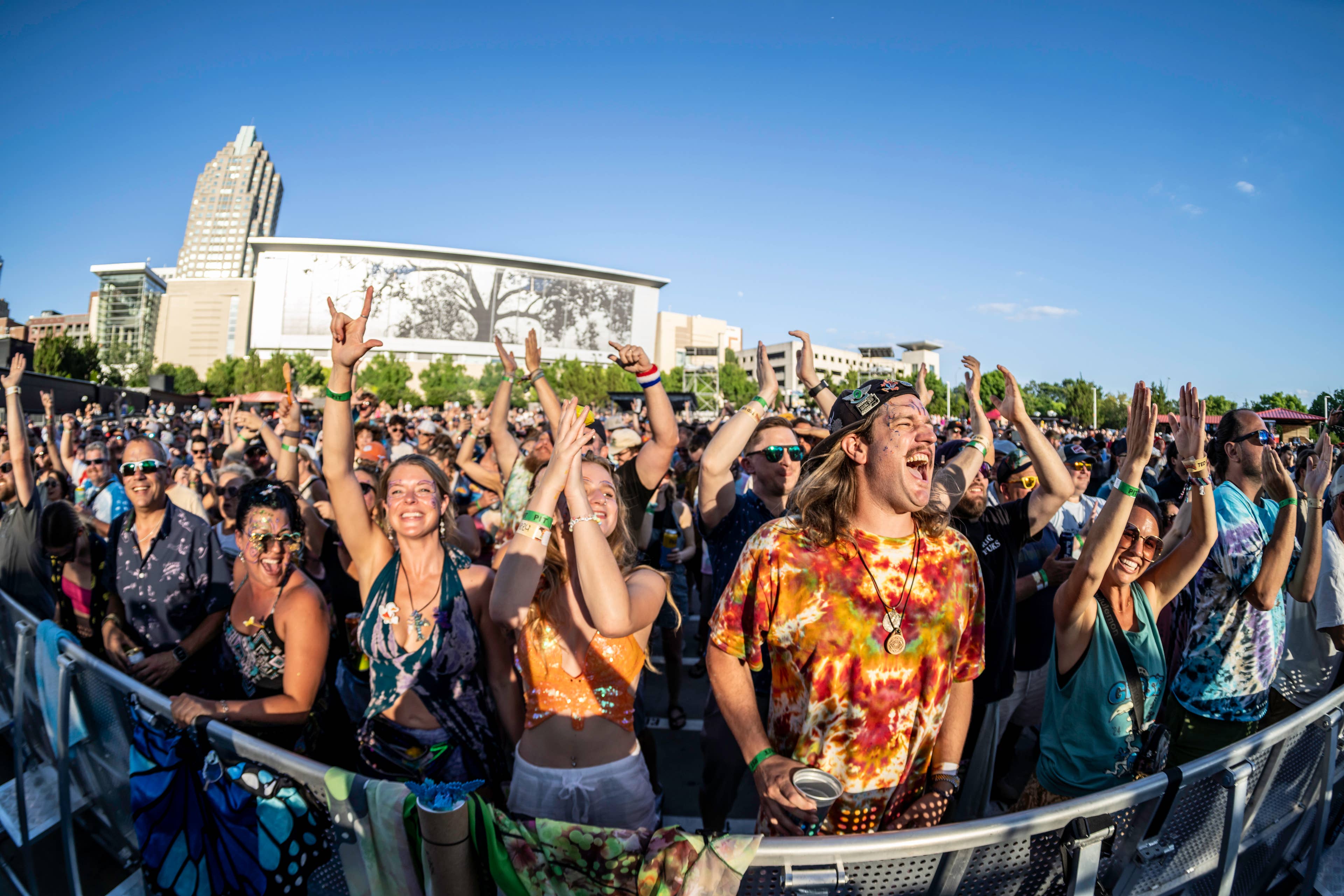 Concert fans in colorful clothes cheer along to a concert on a sunny day with a blue sky.