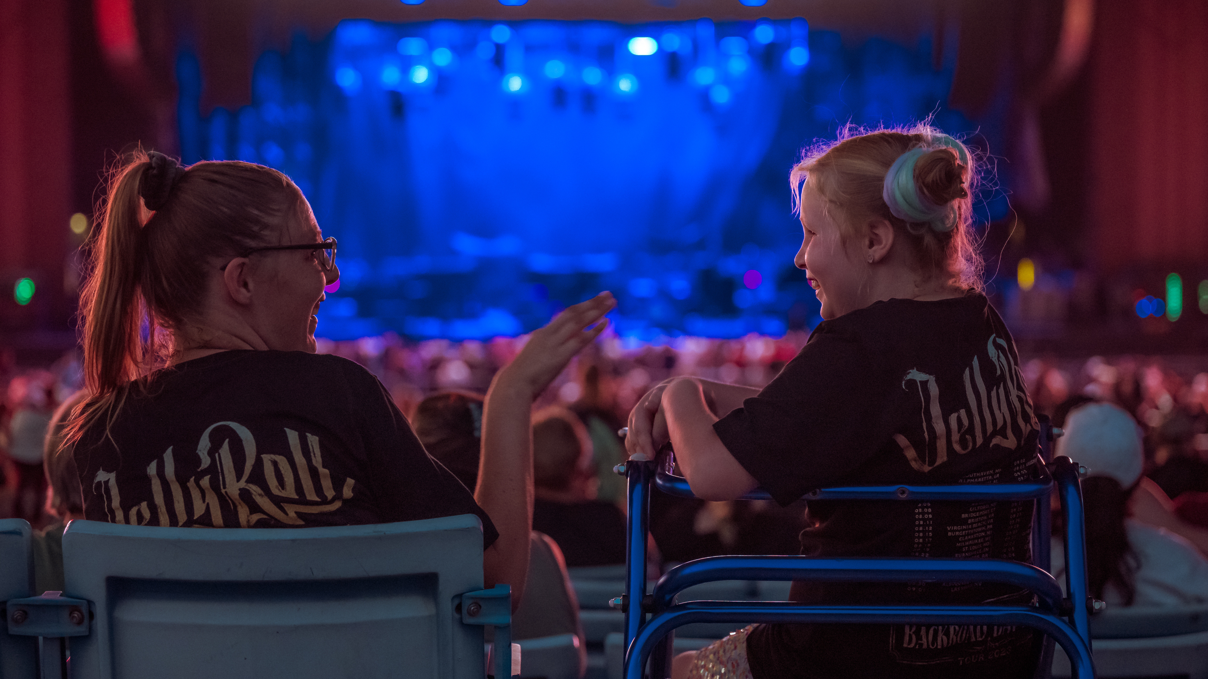 Two fans seated in an accessible section share a moment before a concert, with a large crowd and stage bathed in blue lighting in the background.