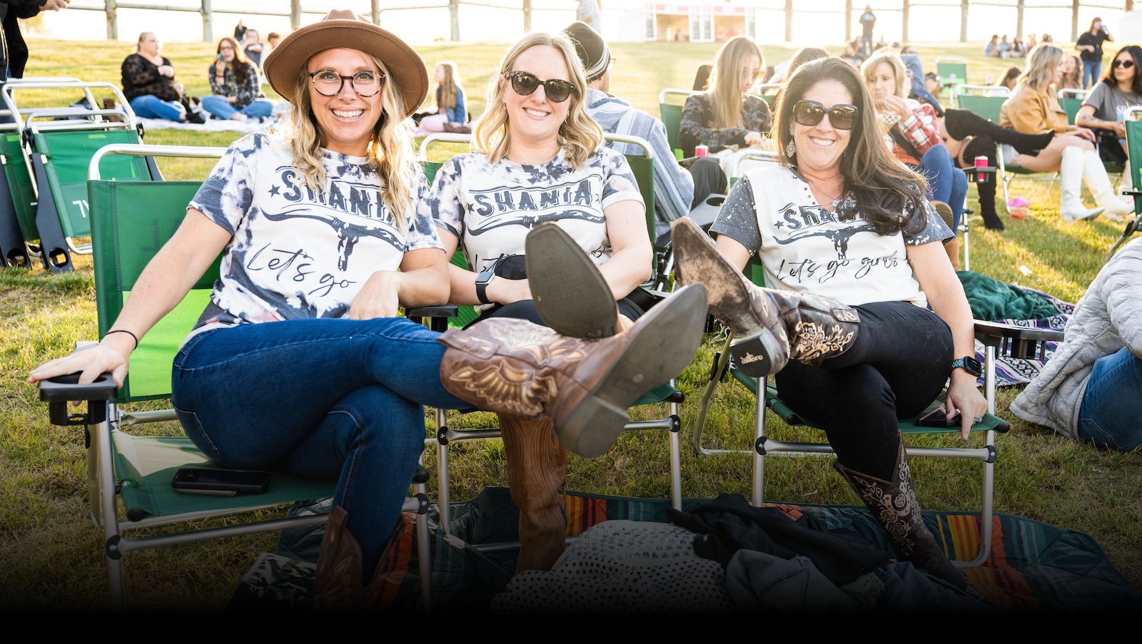 3 people sitting in lawn chairs on the grass, smiling at the camera and showing off their boots