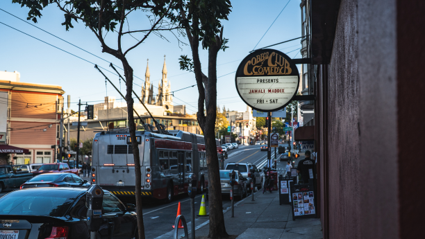 Cobb's Comedy Club marquee displaying an upcoming show, set against a sunset sky, with a bus passing in front of the club entrance.