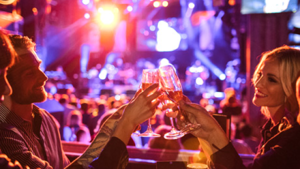 A group of people clinking champagne glasses while sitting in a VIP table with a stage and crowd in the background