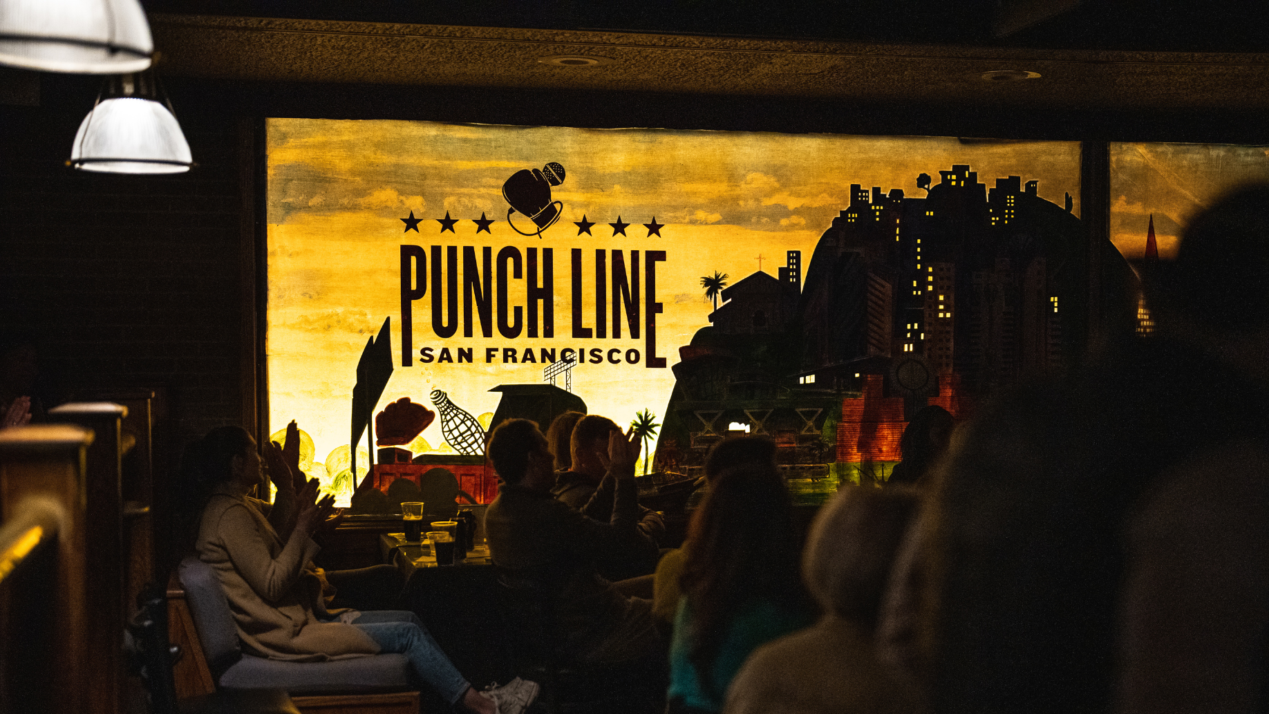 Crowd seated in a dark showroom of Punch Line San Francisco. Part of the club's stage backdrop is displayed behind the crowd. 