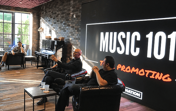 Employees presenting while sitting in chairs on a stage with colorful Music 101 promotional wall featuring text 'Promoting the Next Generation of Live