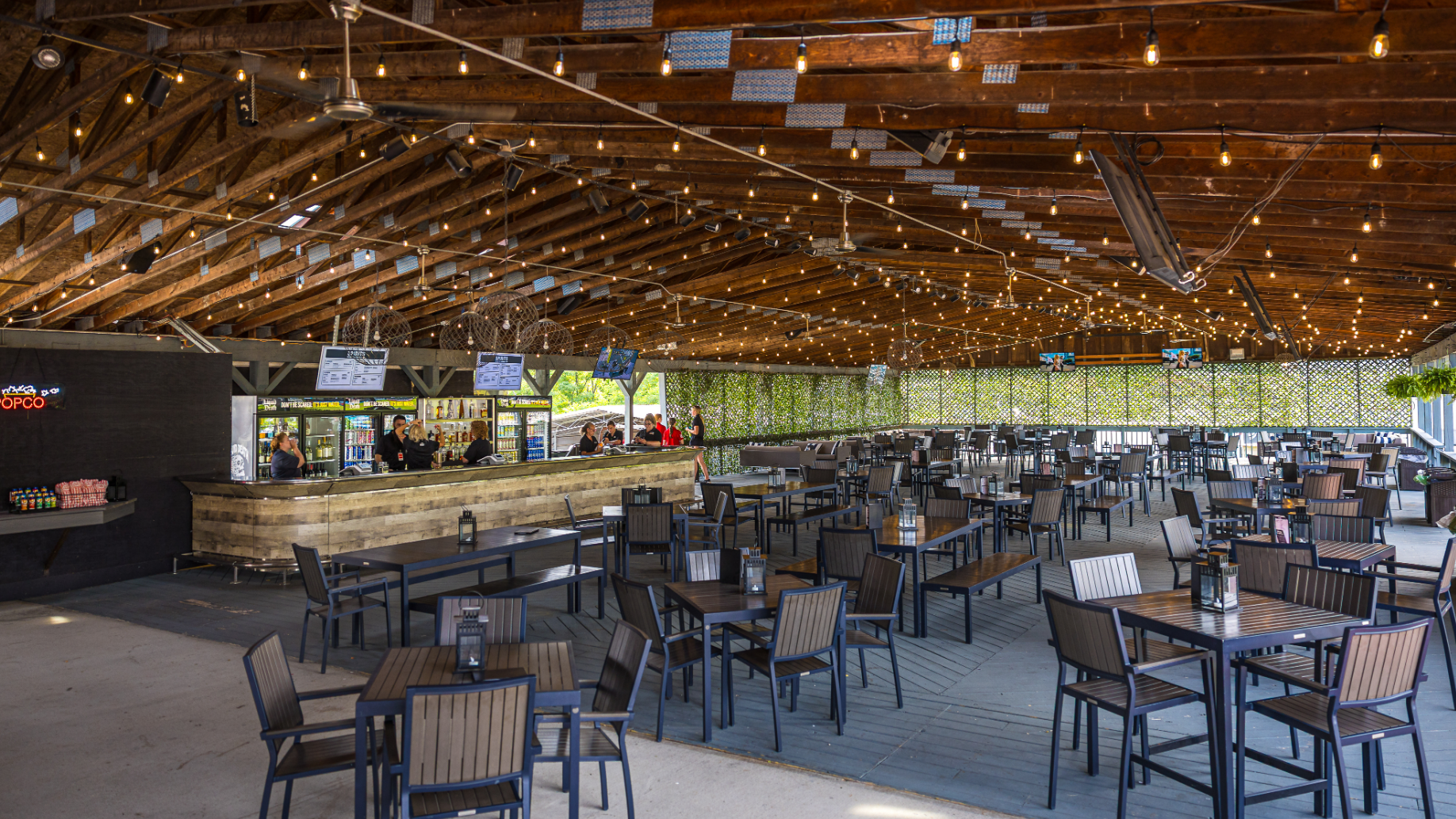 Empty WesBanco lounge showing multiples tables and chairs and employees at a private bar, with string lights hanging from the ceiling. 