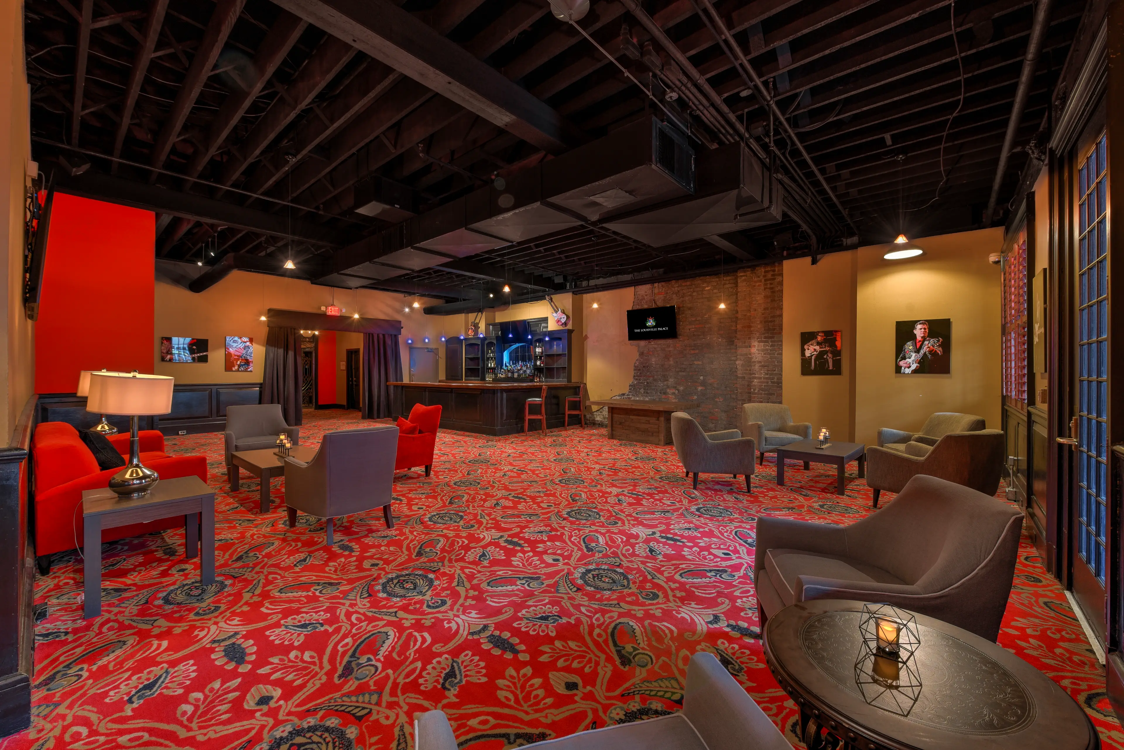 The Marquee Lounge at the Louisville Palace, featuring plush chairs, warm lighting, exposed brick and beams, a full-service bar, and bold red patterned carpet for a stylish yet relaxed vibe.