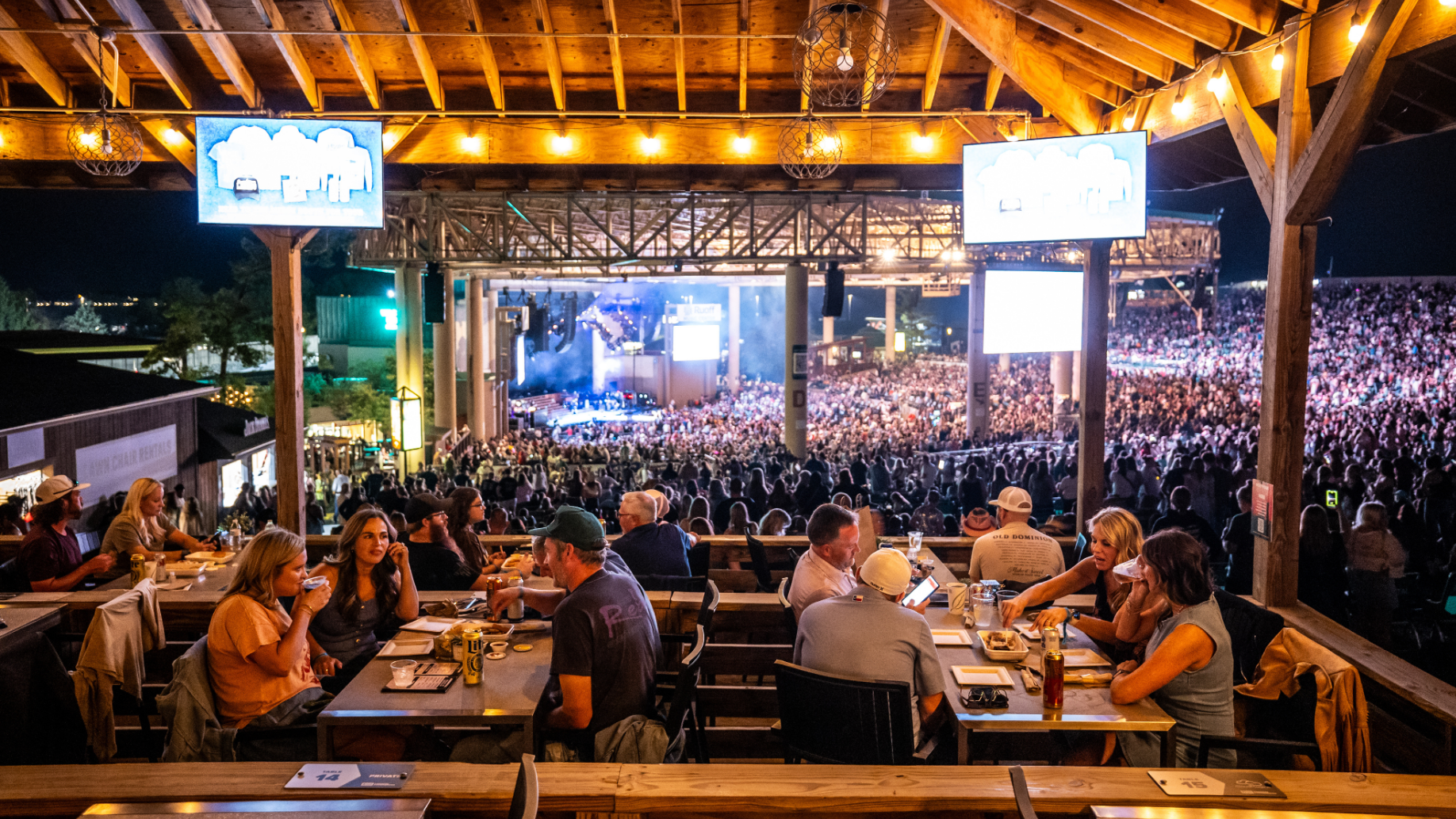 Fans eating and drinking at tables on a platform with multiple decks, overlooking other fans on the lawn and in the concourse with the concert stage in the far background. 