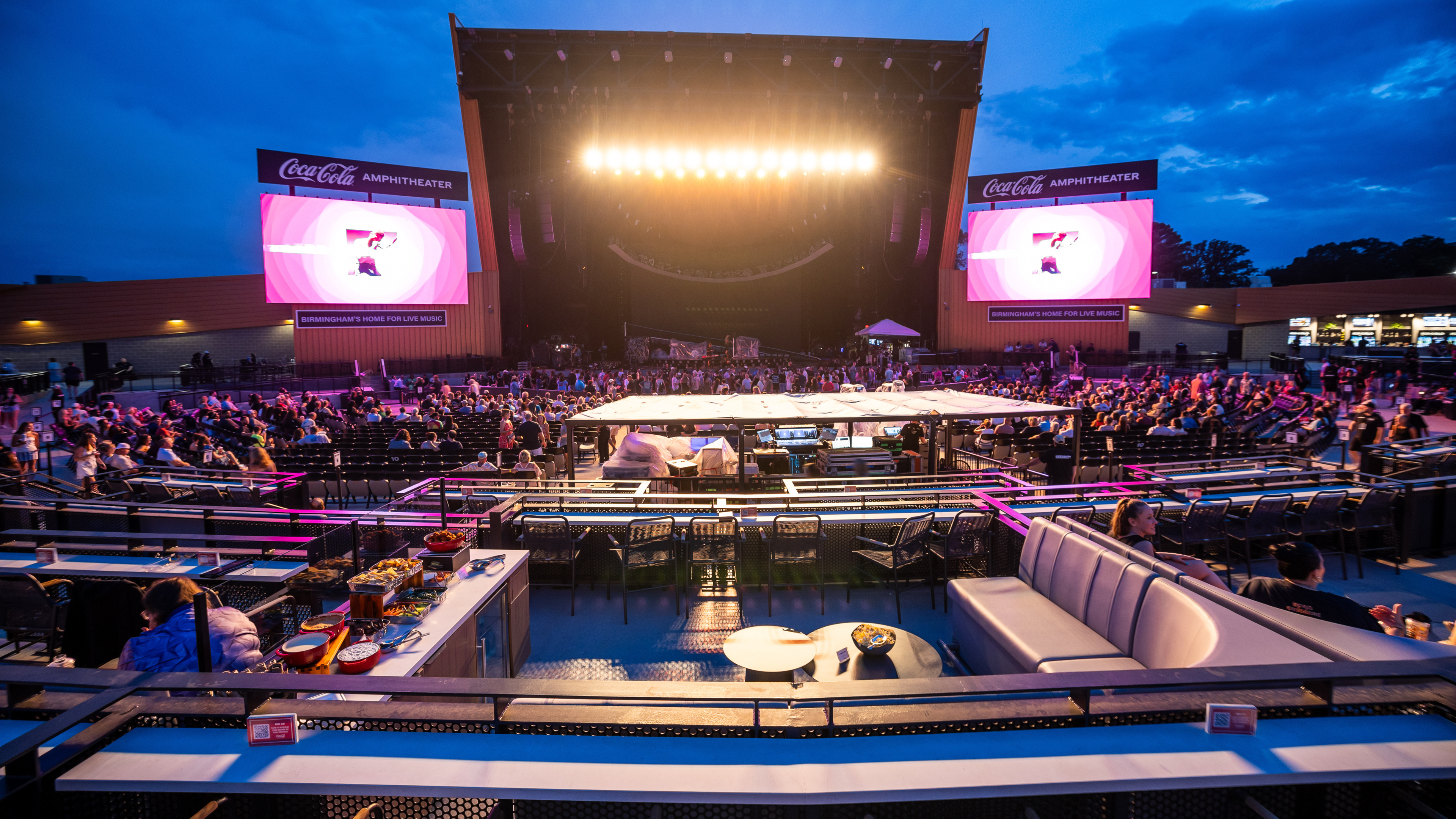 Wide view of an outdoor concert amphitheater at dusk before a performance, with a brightly lit stage, two large video screens, audience seating filling with people, and a VIP lounge with couches and refreshments.