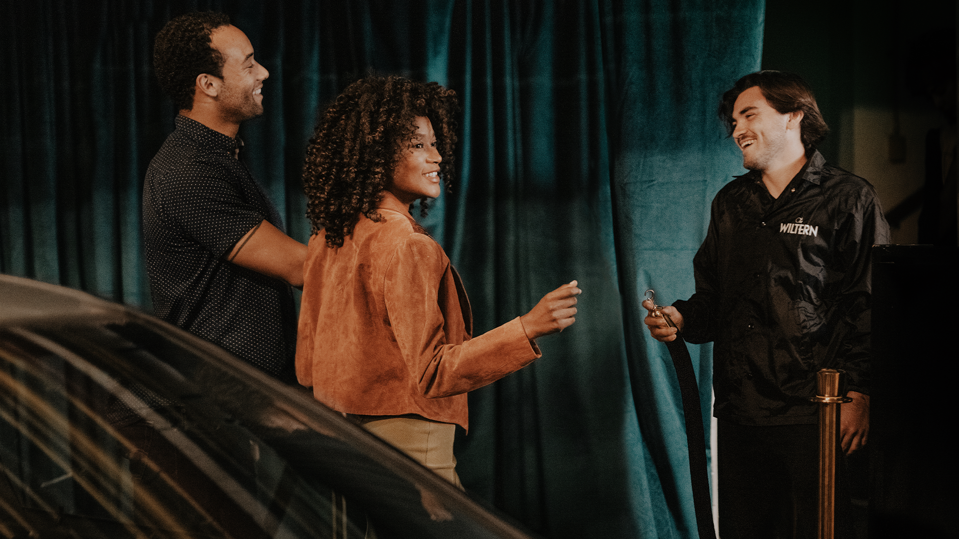 Three people engaging in a friendly conversation beside a car, with one person holding car keys. They are in a showroom-like setting with a dark blue curtain backdrop.