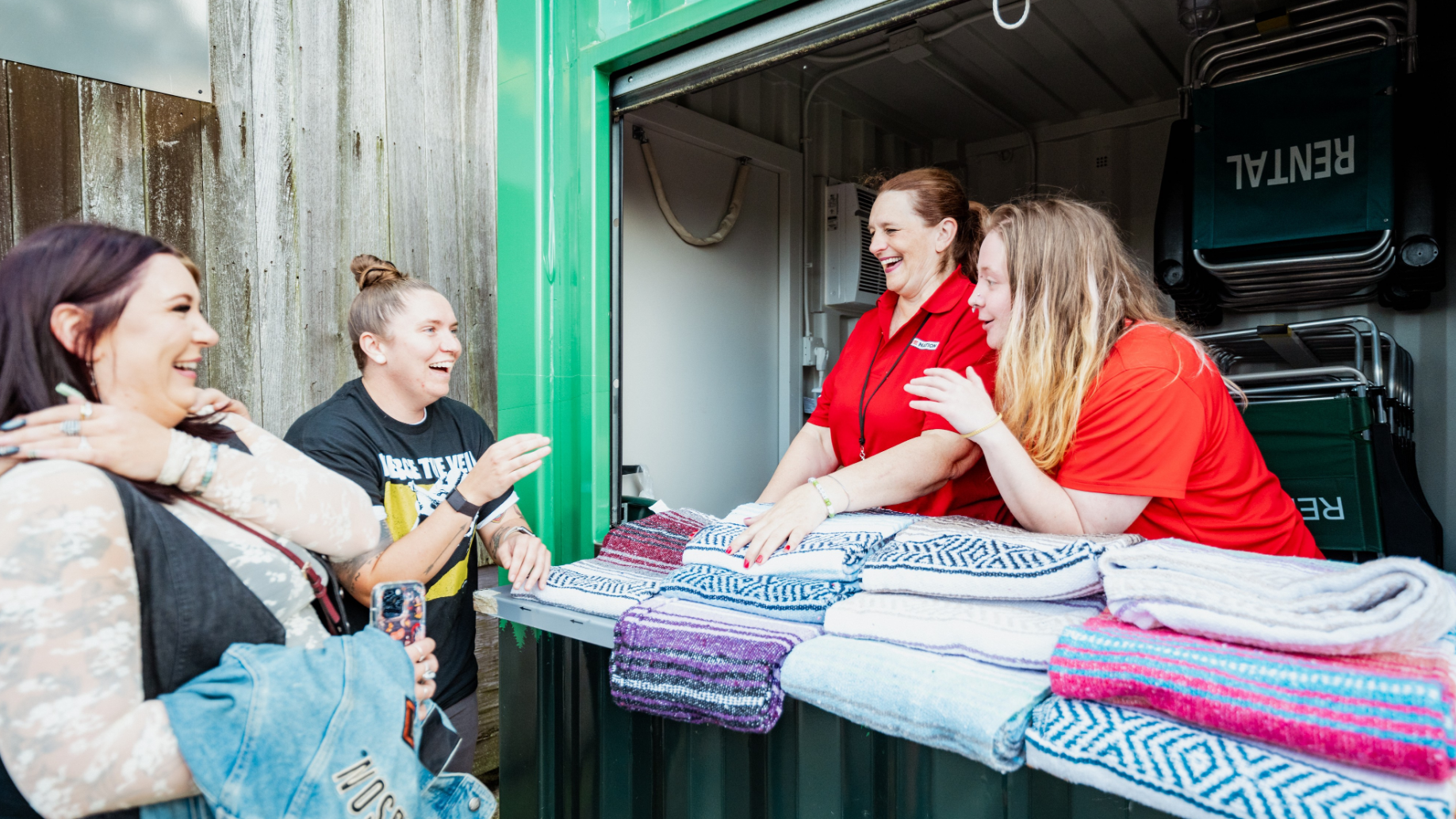 Two employees smiling and talking with fans at the blanket rental stand. Multiple blankets on display in front of them. 