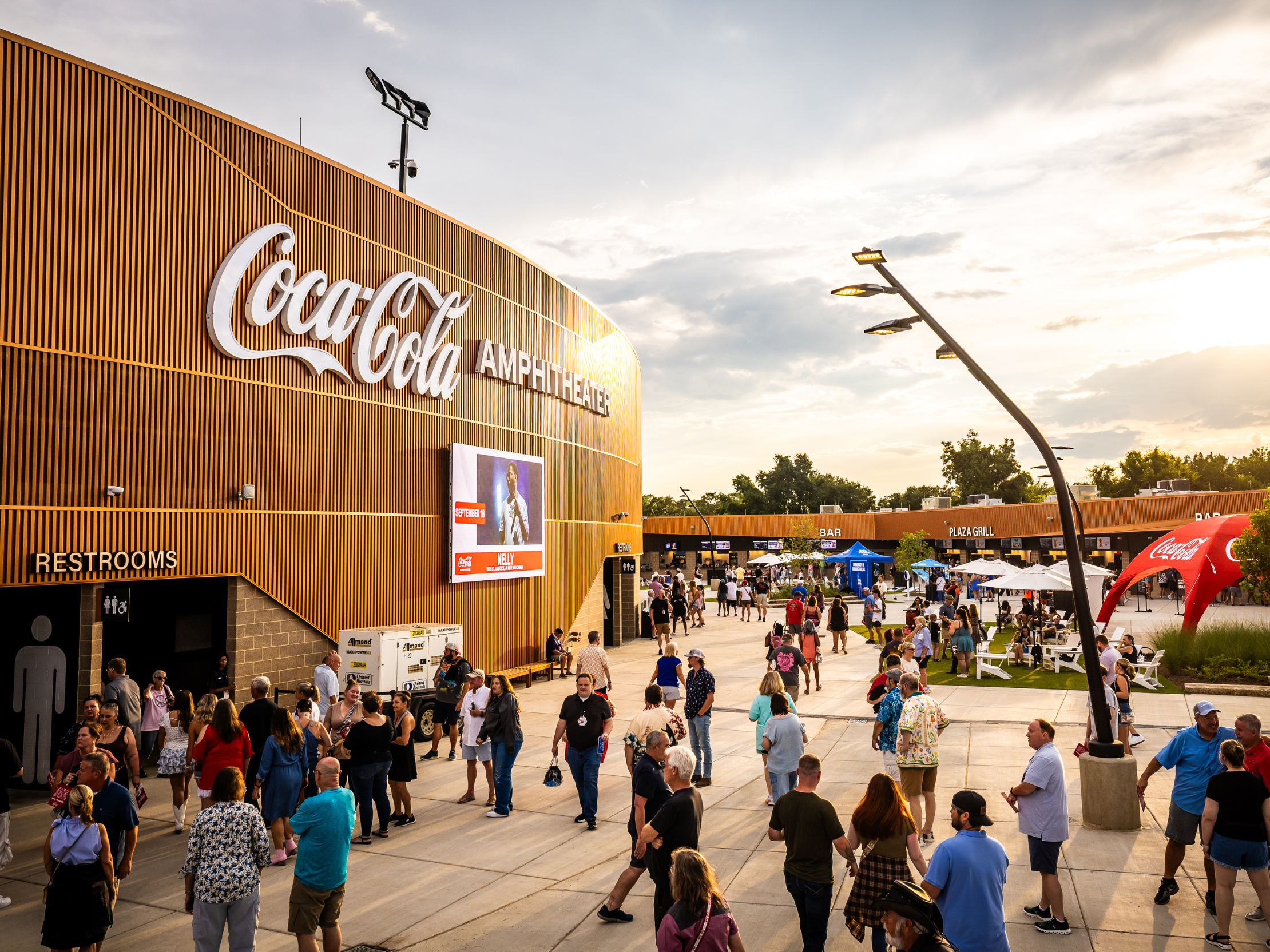 Crowd arriving at Coca-Cola Amphitheater entrance plaza at sunset with venue signage visible.