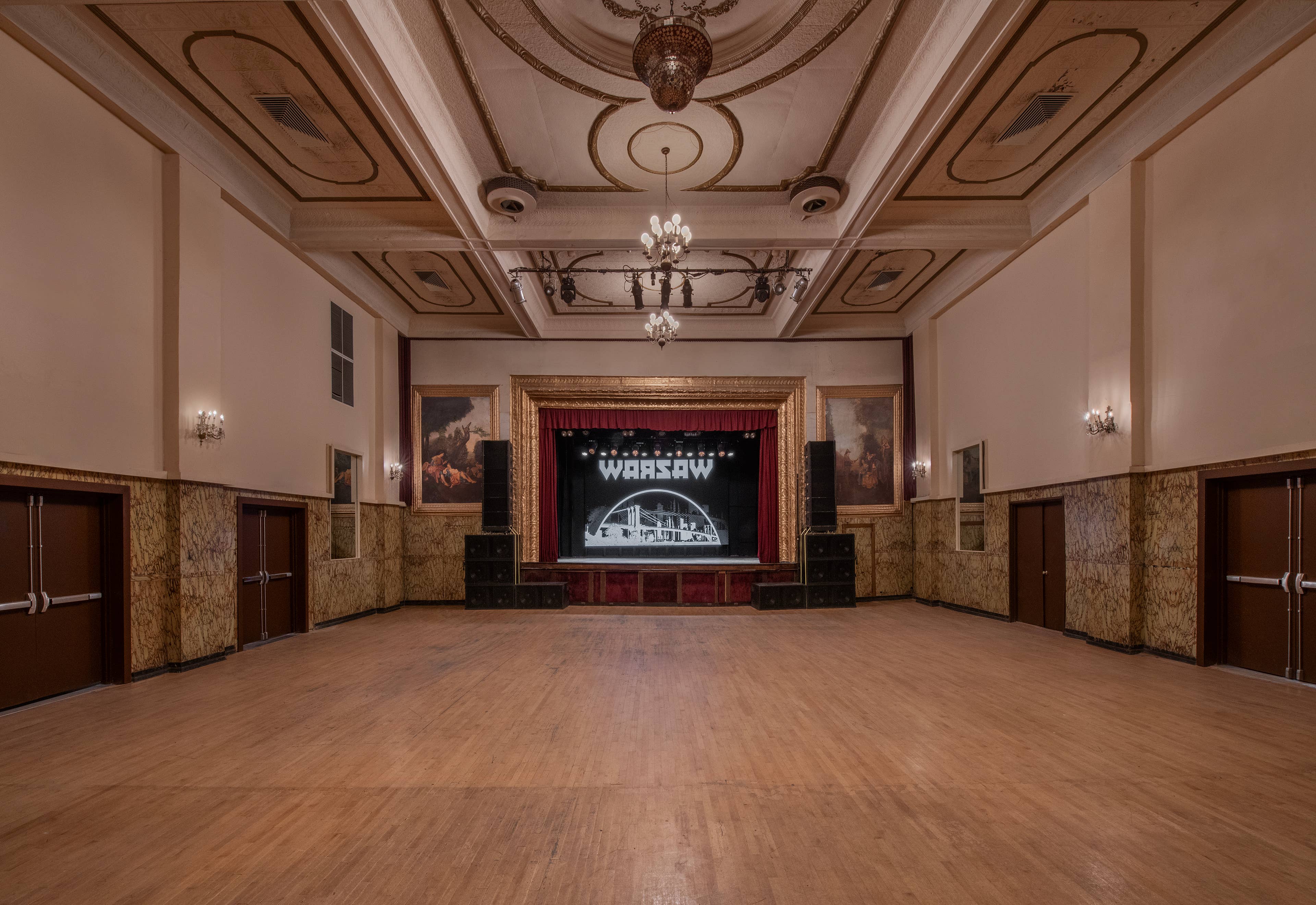A photo of Warsaw's main ballroom showing the stage with a gold frame around it and large paintings on both sides along with the beautiful ceiling details