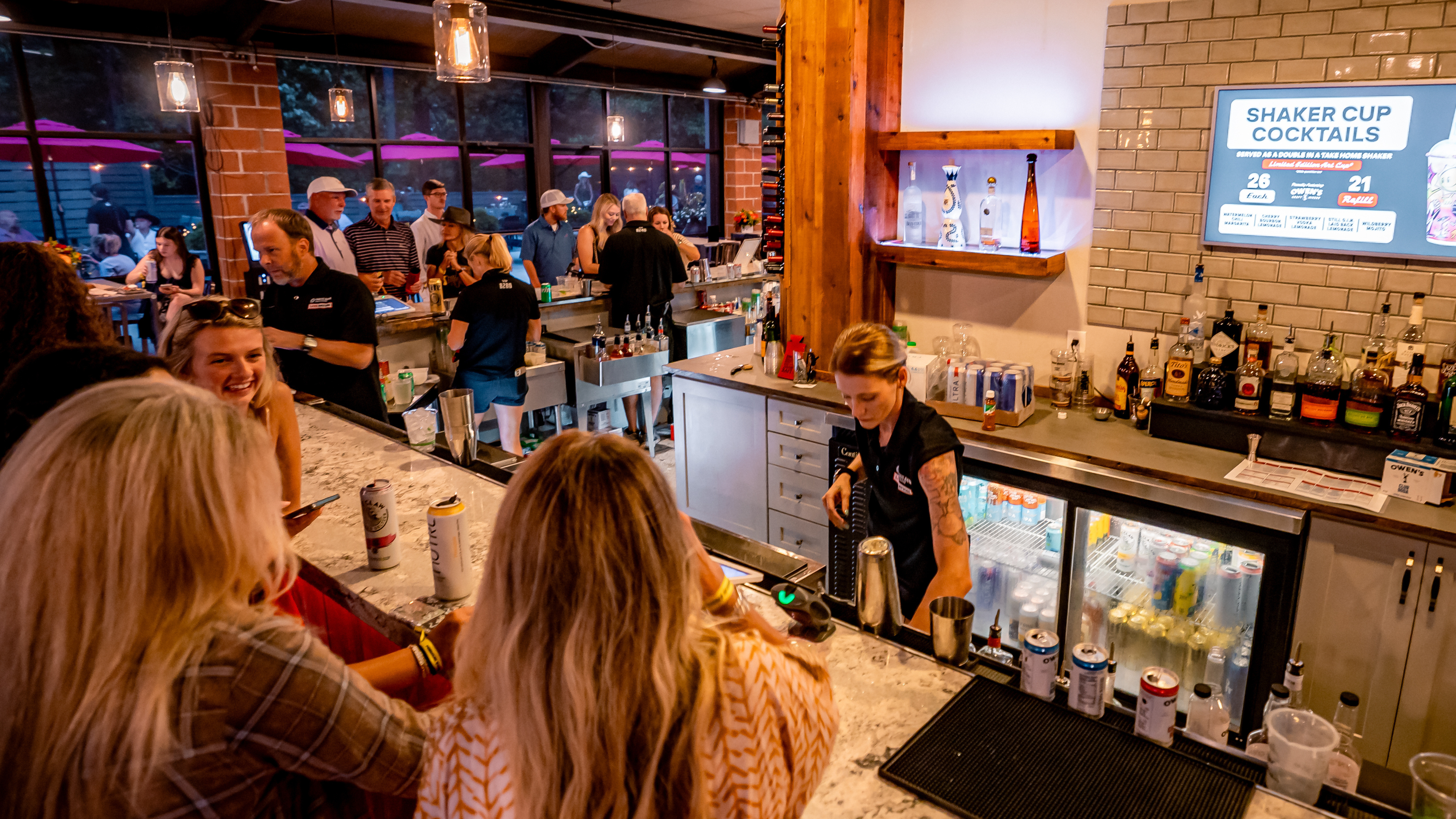 Indoor concert lounge bar with guests ordering drinks at a full-service counter, staff serving cocktails, and a backlit spirits display, plus an outdoor patio in the back with bench seating.