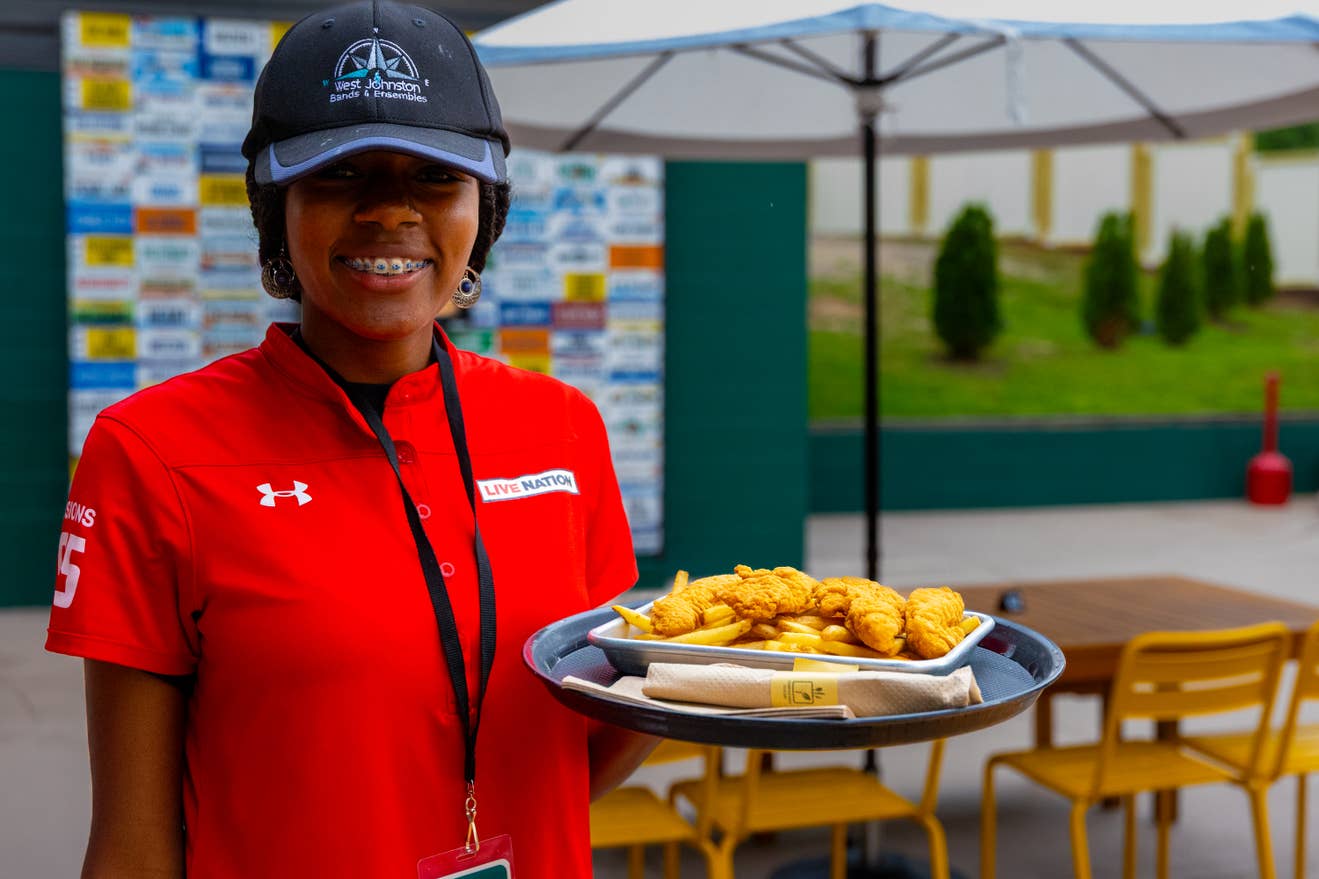 A female venue worker in a red uniform shirt smiles while holding a plate of chicken tenders.