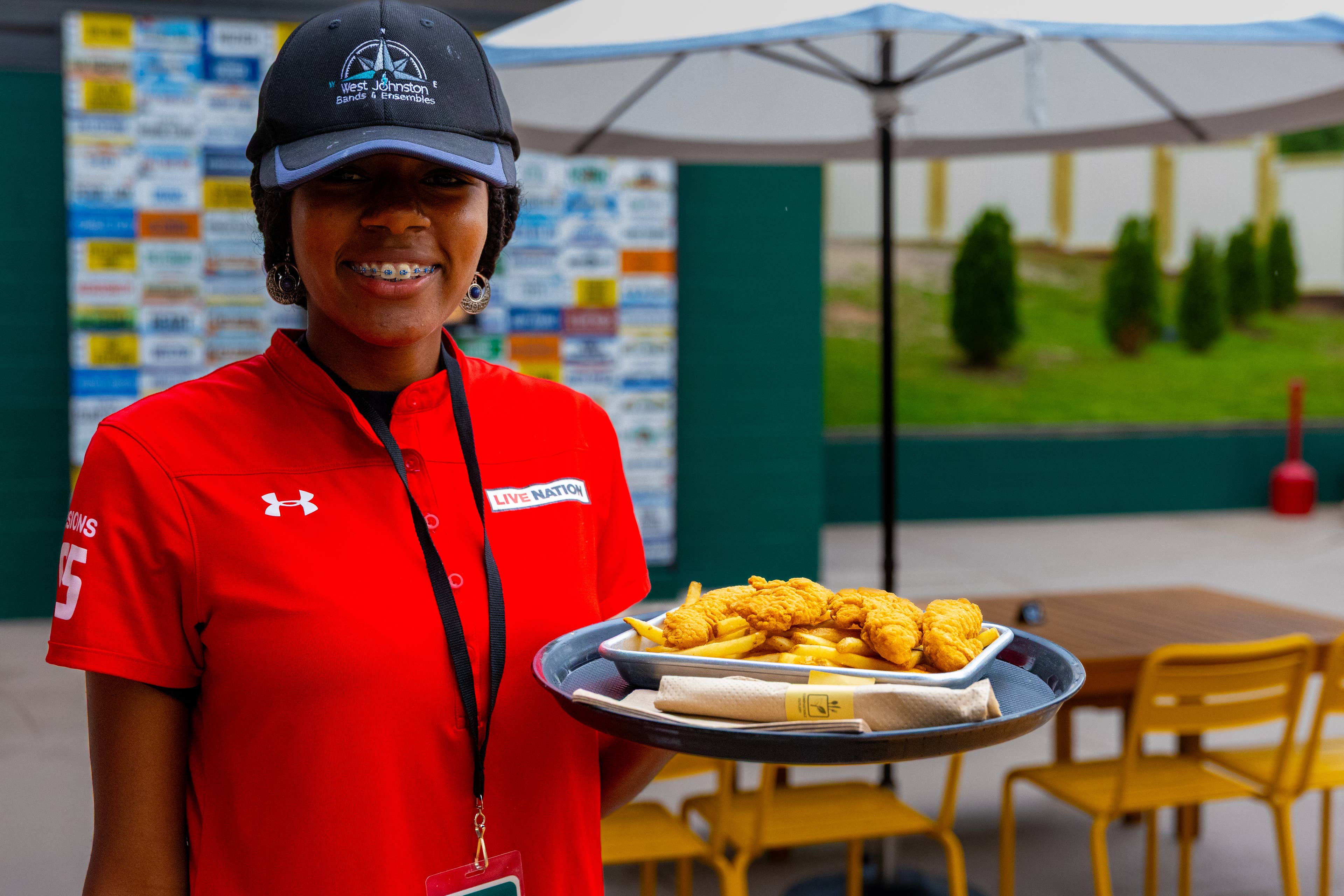 A female venue worker in a red uniform shirt smiles while holding a plate of chicken tenders.