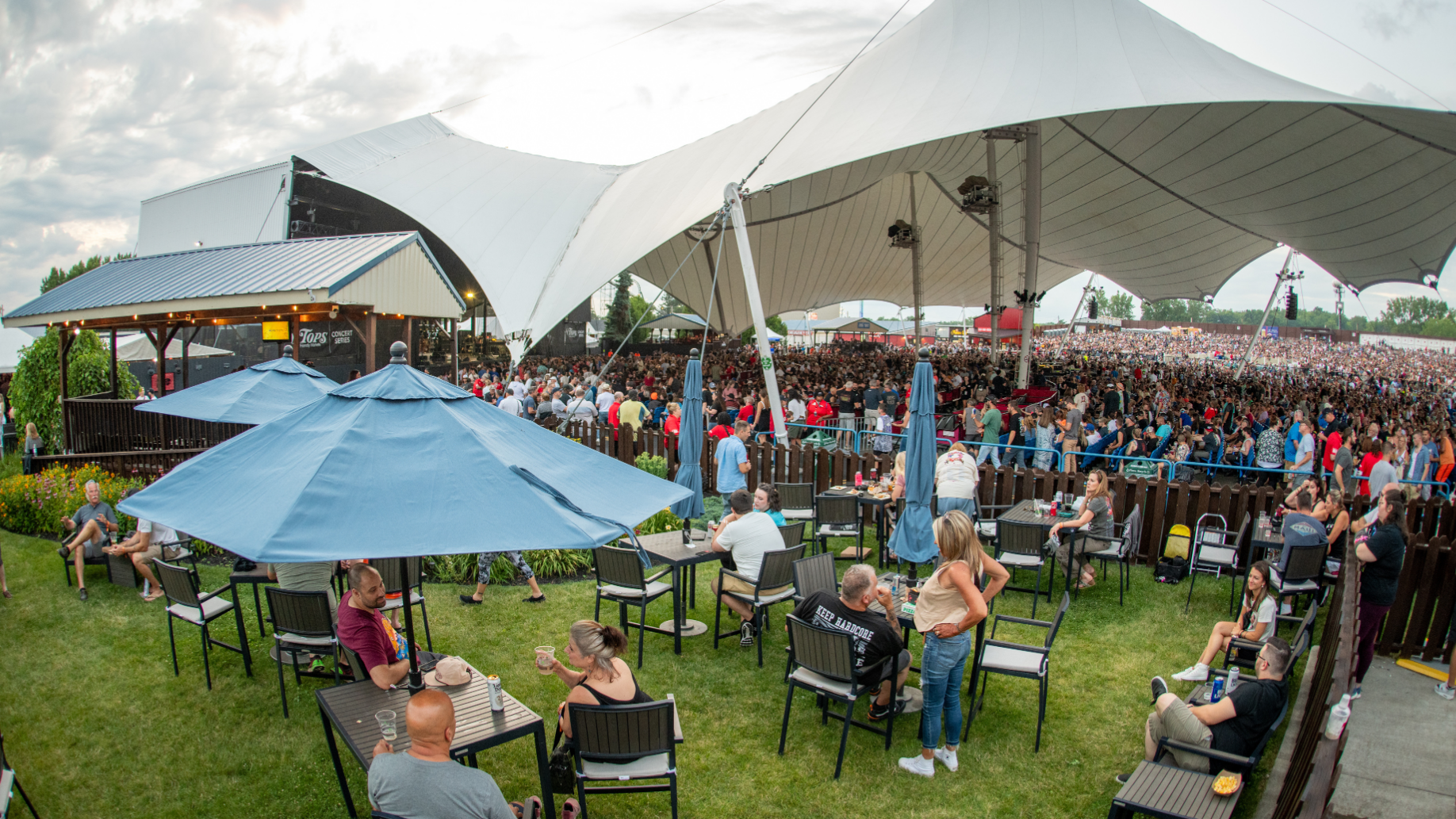 Fans sit around tables in a grassy area to the left of the concourse, shaded by large blue umbrellas.