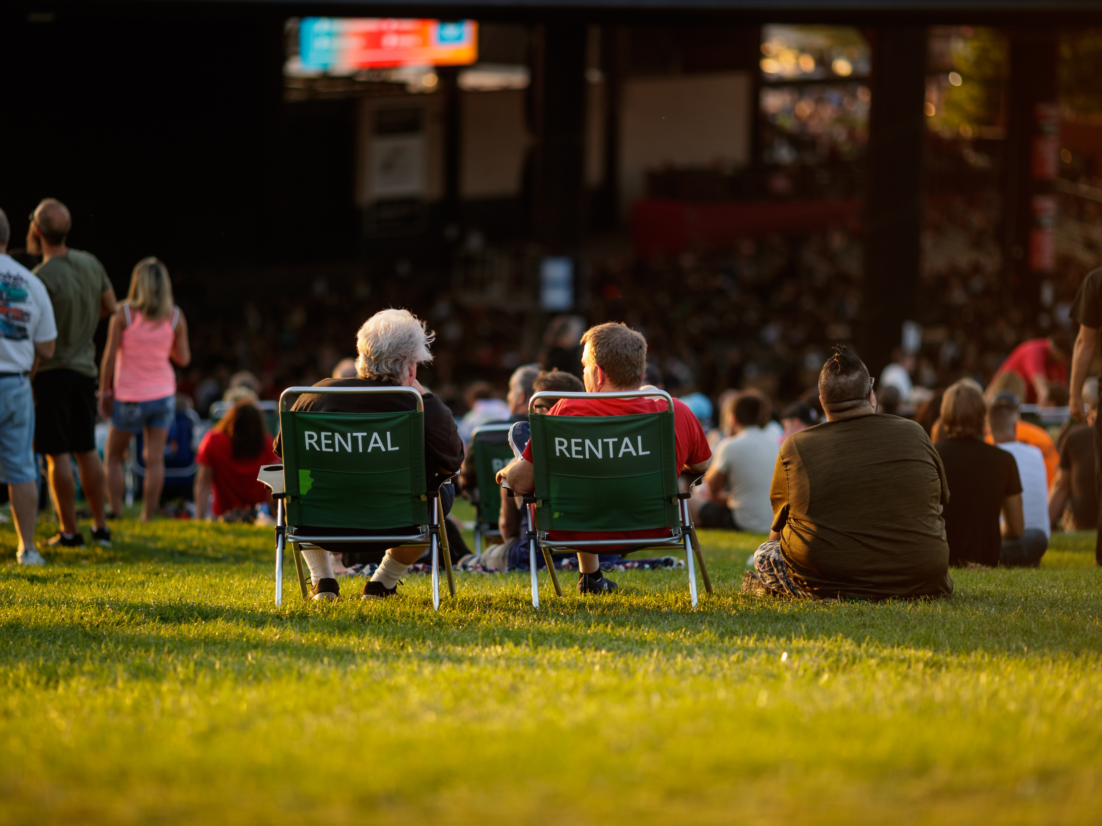 Three fans sitting in the Lawn at Credit Union 1 Amphitheatre. Two are using the Rental Lawn Chairs, one is on the ground. 