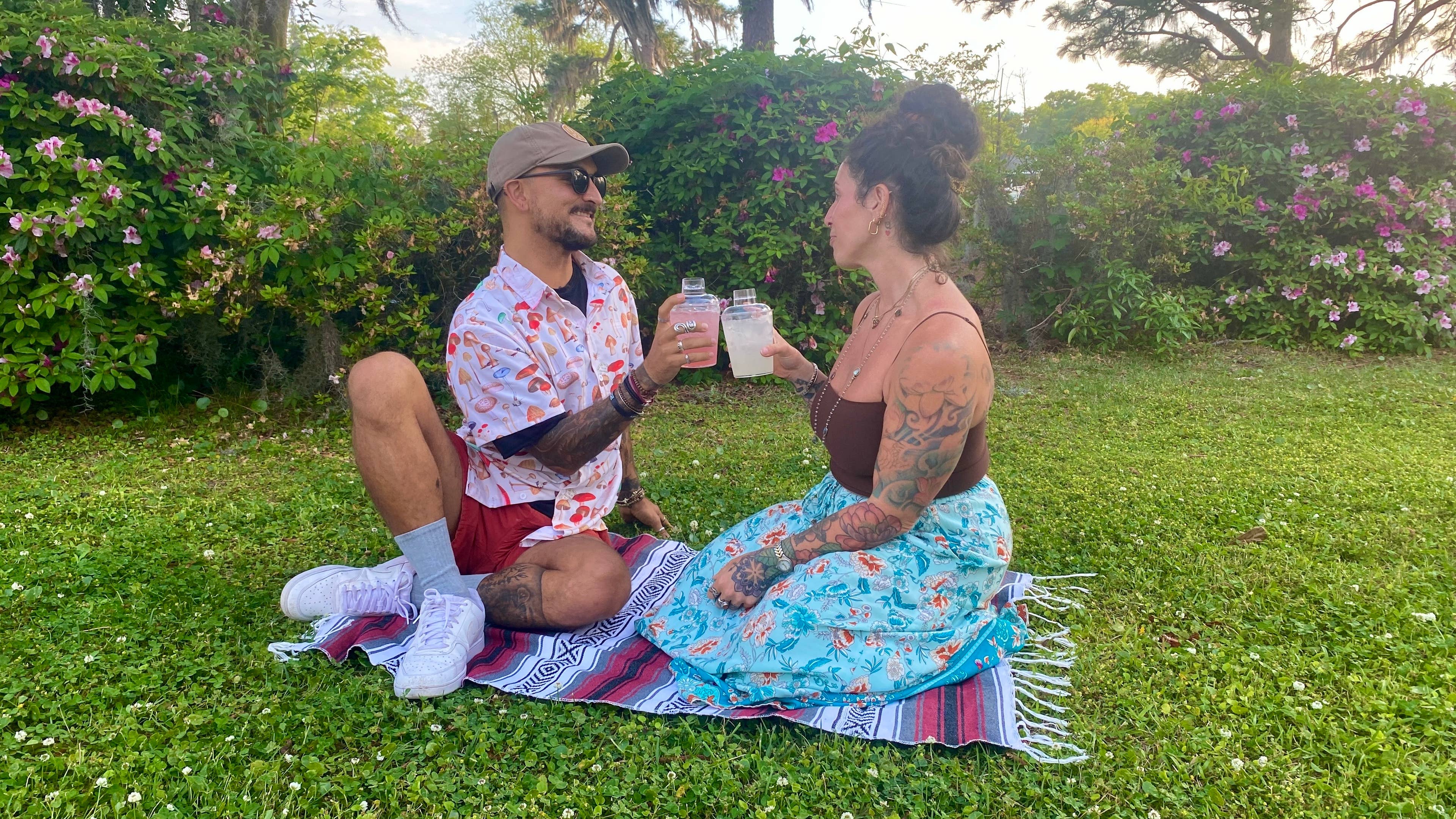 A man and a woman sitting on a blanket in the lawn with drinks.