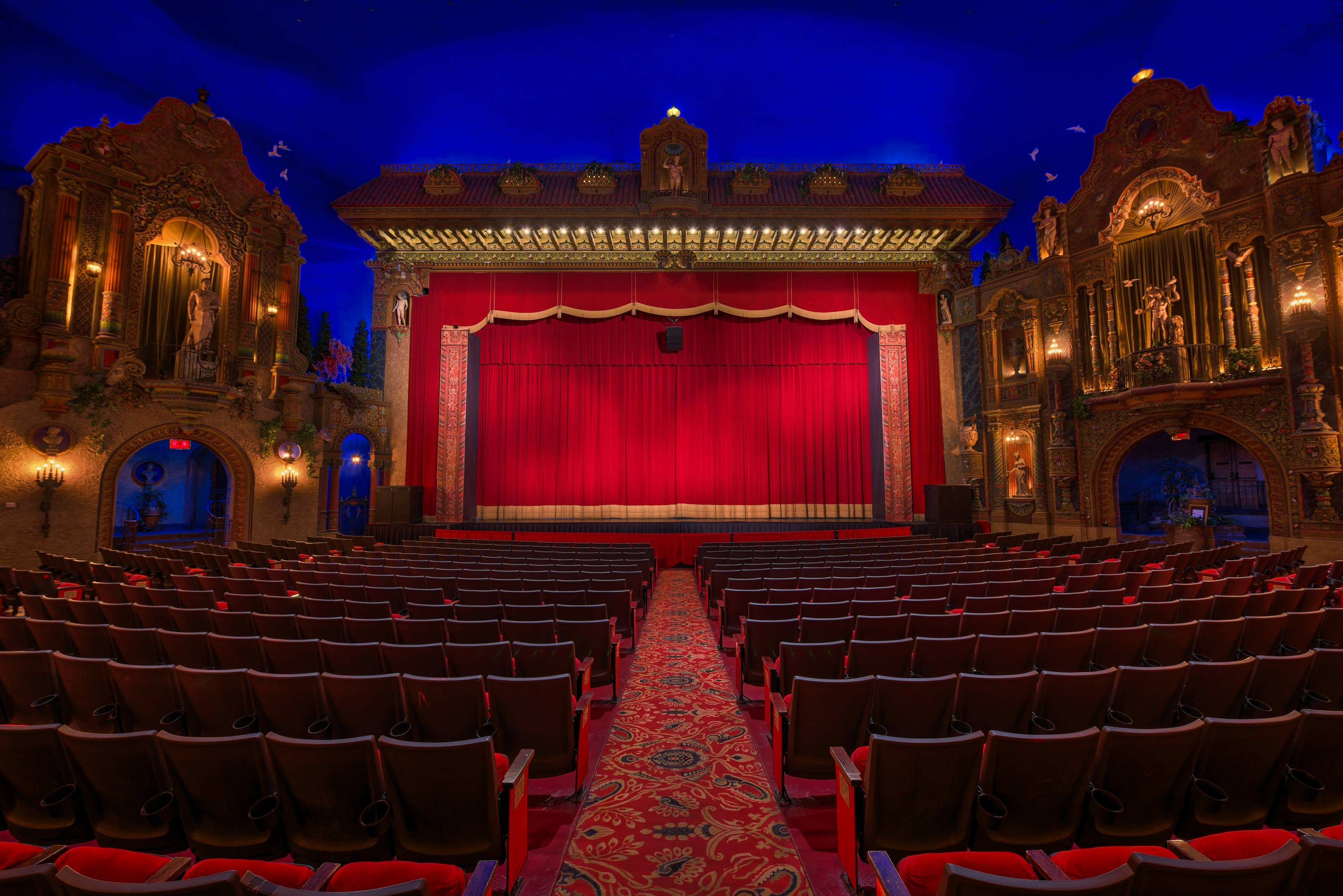 The theatre stage surrounded with ornate architecture, red velvet seating, and red curtains.