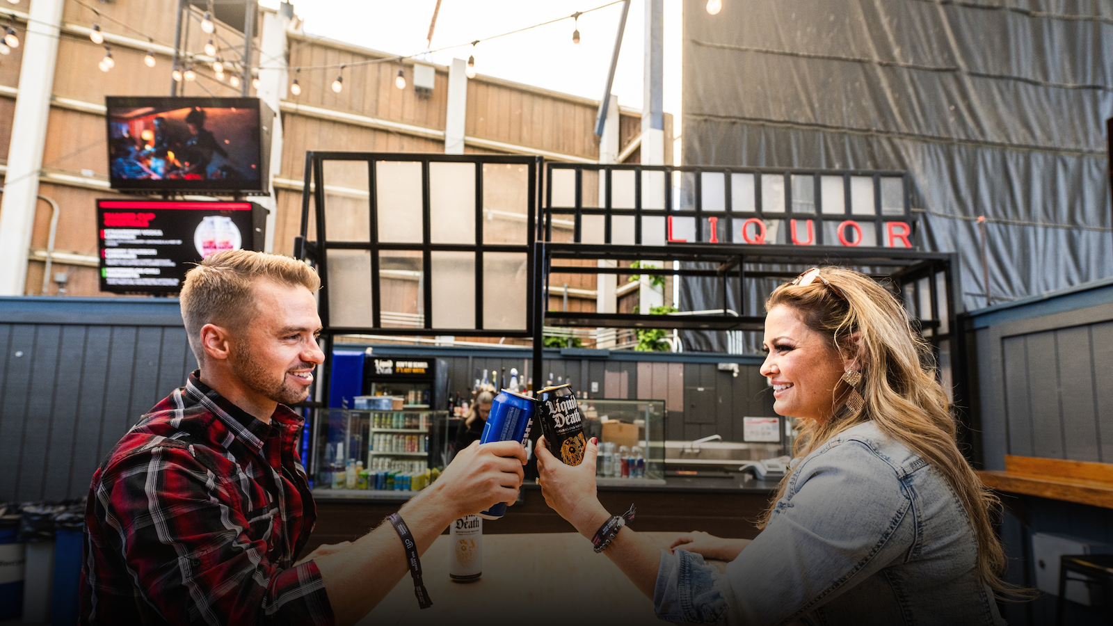 Two people are shown smiling at each other and clinking their drinks, with the bar in the background. Seated at a wooden table with the venue in the background.