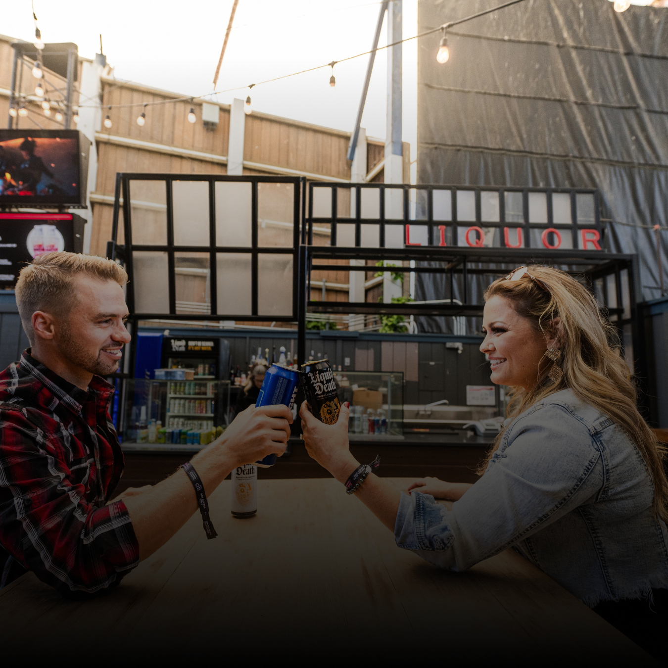 Two people sitting at a table in front of a bar, cheersing their drinks and smiling