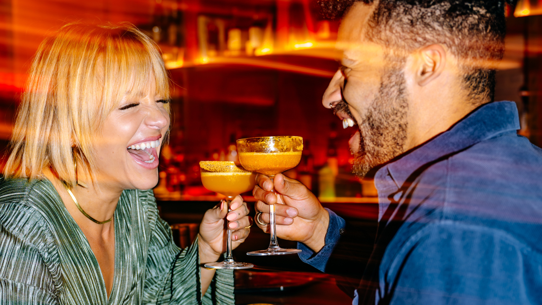 A girl and a guy cheers orange cocktails and laugh while sitting at the bar.