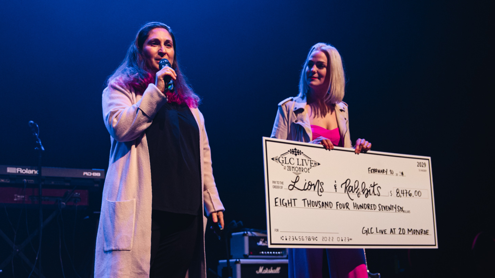 A woman speaking into a microphone and a lady standing next to her holding a giant check to giveaway.