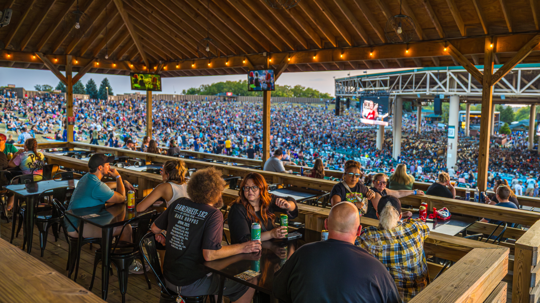 Fans drinking at tables on a platform with multiple decks, overlooking other fans on the lawn and in the concourse. String lights hang from the platform roof.