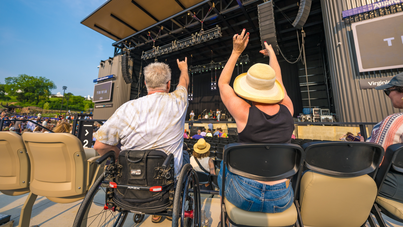 Two adults raise the rock sign with their fans from their chairs, one in a wheelchair, as they watch a concert.