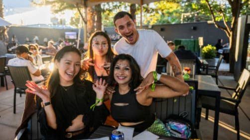 People are shown seated at a table under a large tent, with food and drinks in front of them. They have big smiles on their faces and fun makeup on.